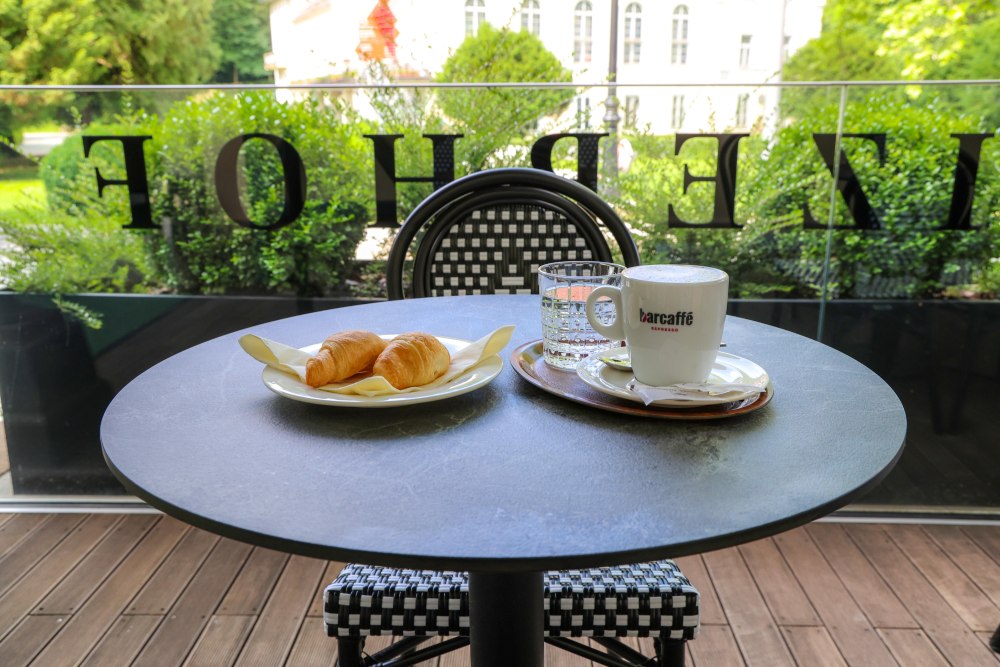 Ein Tisch mit zwei Croissants, einer Tasse Kaffee und Wasser. Im Hintergrund grüne Pflanzen und ein schönes Gebäude.