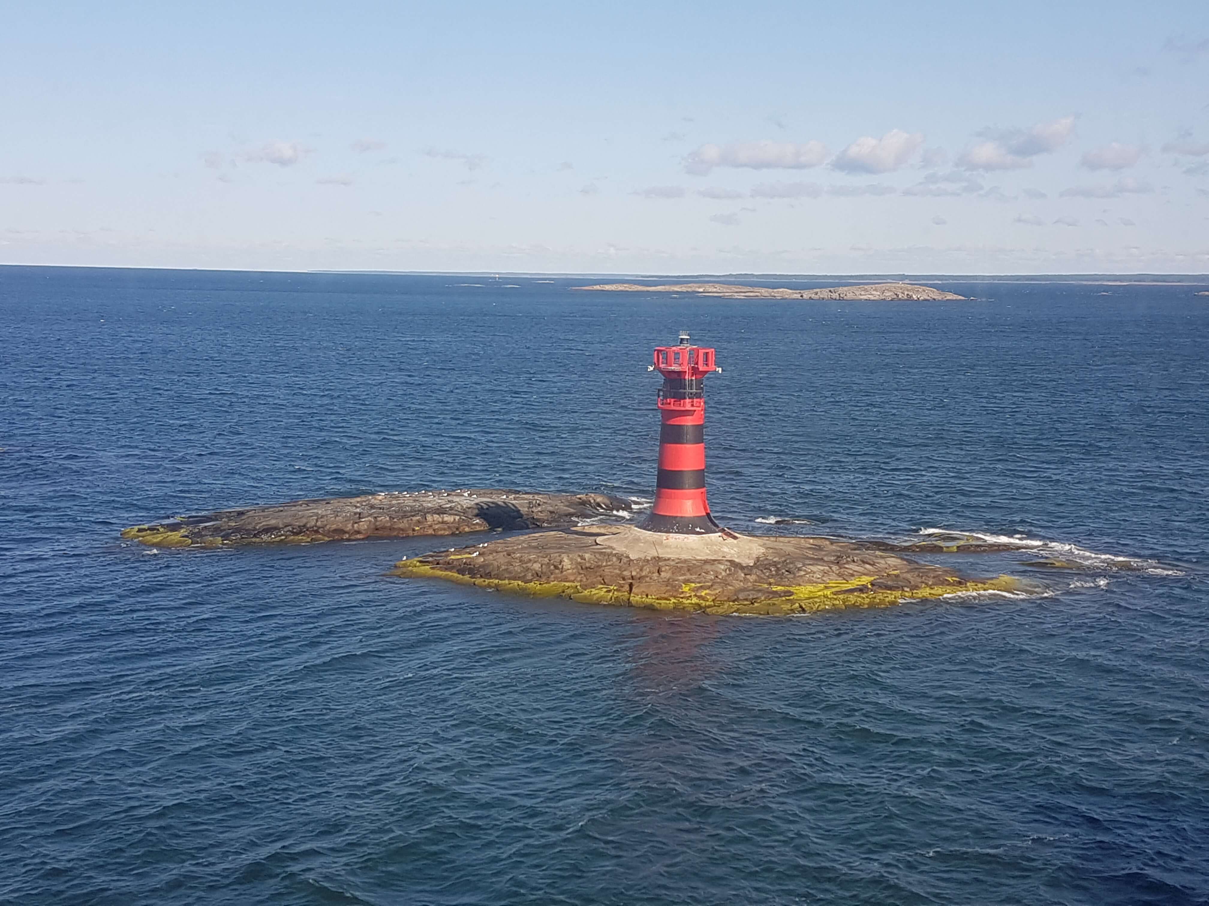 Ein rot-schwarz gestreifter Leuchtturm steht auf einem kleinen Felsen im blauen Wasser. Wolken am Himmel.