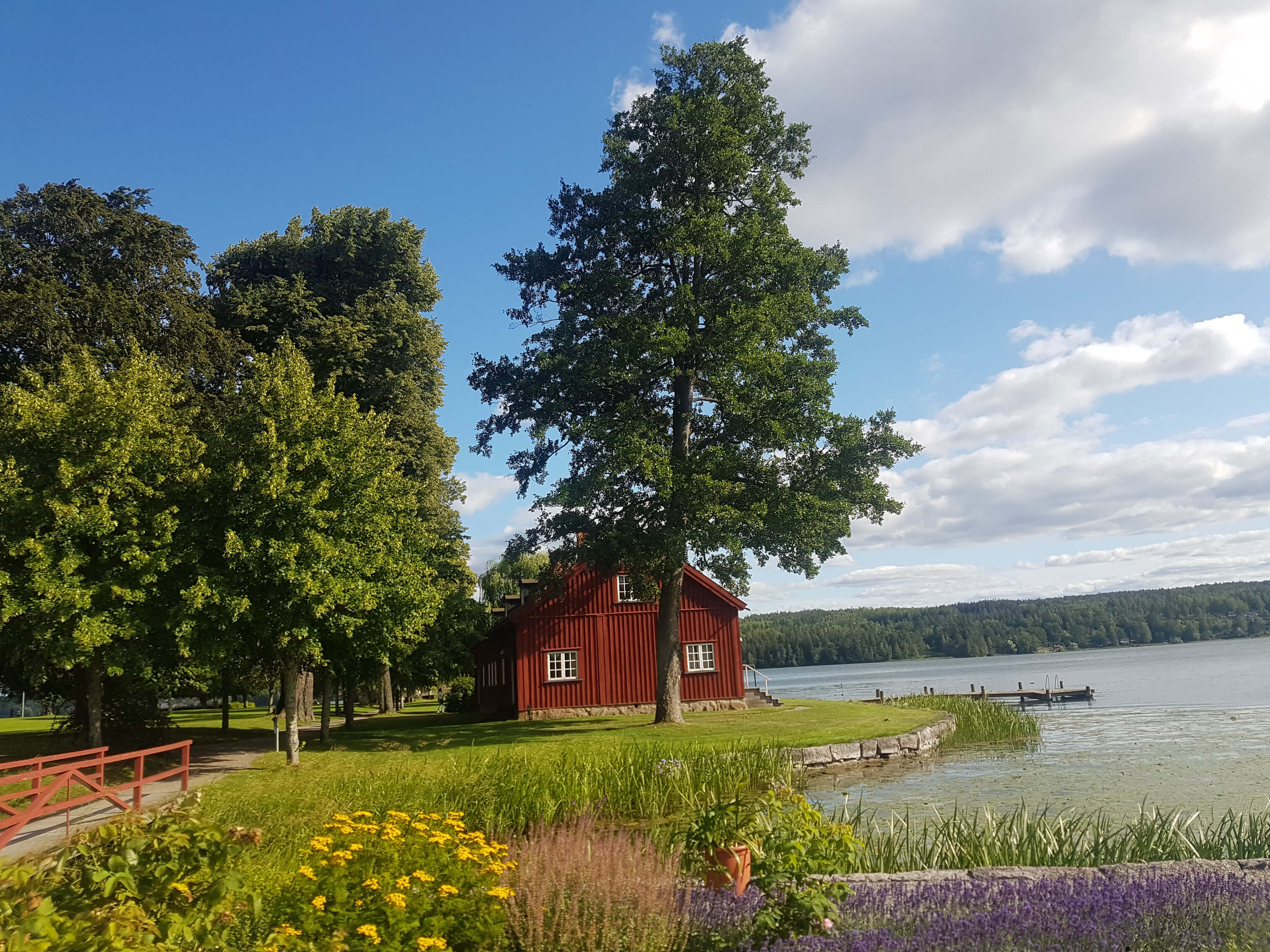 Ruhige Landschaft mit einem roten Holzhaus, Bäumen, Blumen und einem ruhigen See unter blauem Himmel.