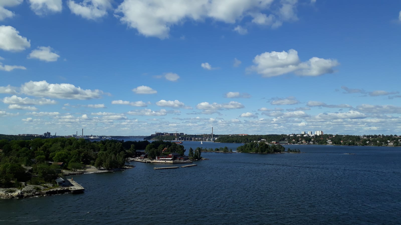 Ein malerischer Blick auf einen sonnigen See, umgeben von Bäumen und Gebäuden unter einem blauen Himmel mit Wolken.