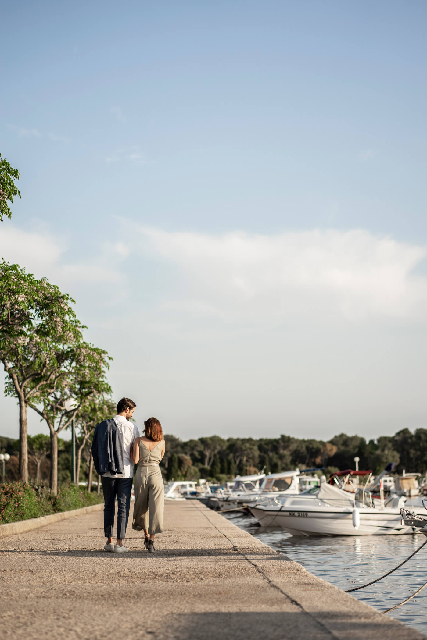 Ein Paar spaziert am Hafen entlang, umgeben von Booten und Bäumen, unter einem blauen Himmel.