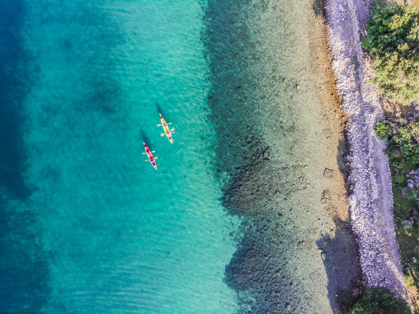 Zwei Kajaks gleiten über kristallklares Wasser an einem steinigen Ufer entlang. Ein schöner sonniger Tag.