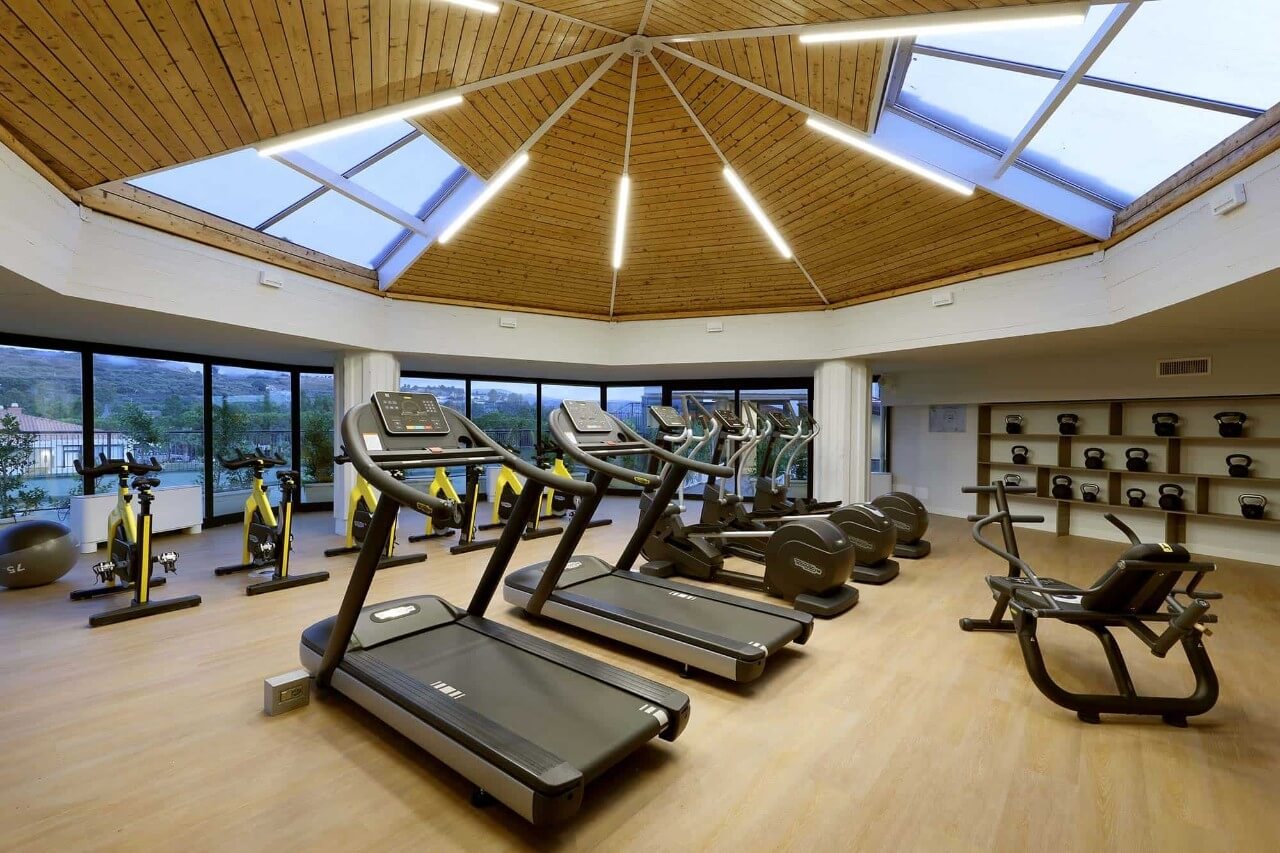 Moderne Fitnessstudio mit Holzbalkendecke, Laufbändern, Fahrrädern und Gewichten, große Fenster für Tageslicht.