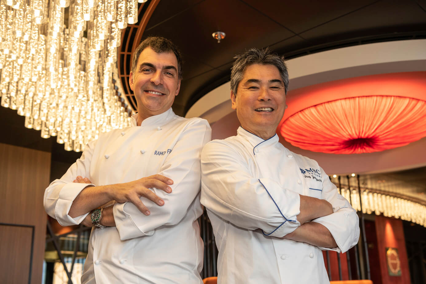 Zwei Köche lächeln in einer eleganten Umgebung unter einer beeindruckenden Deckenleuchte.