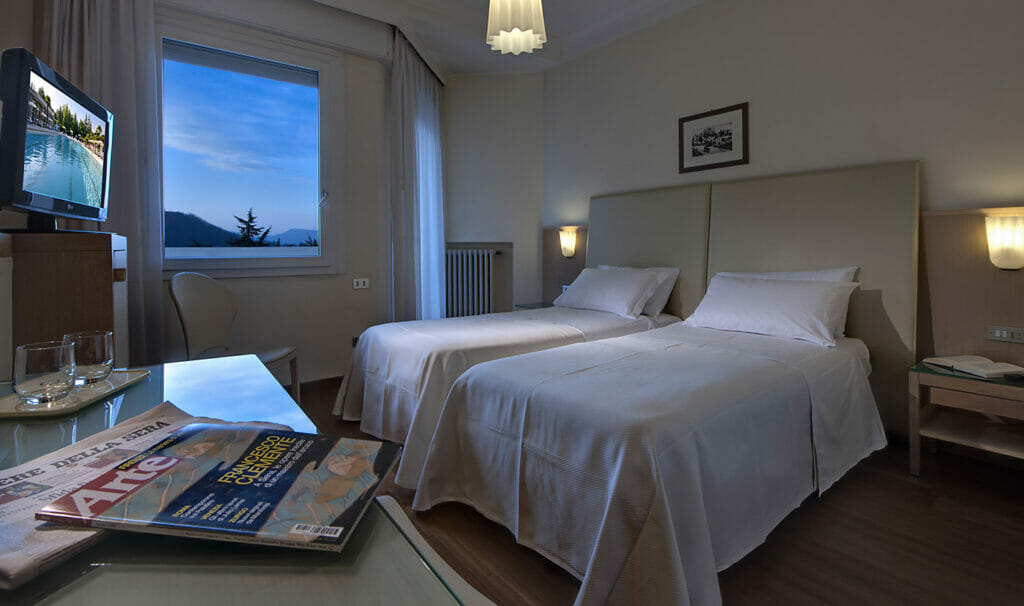 Helles Hotelzimmer mit zwei Betten, Fernseher, Tisch und Blick auf die dämmernde Landschaft durch ein Fenster.
