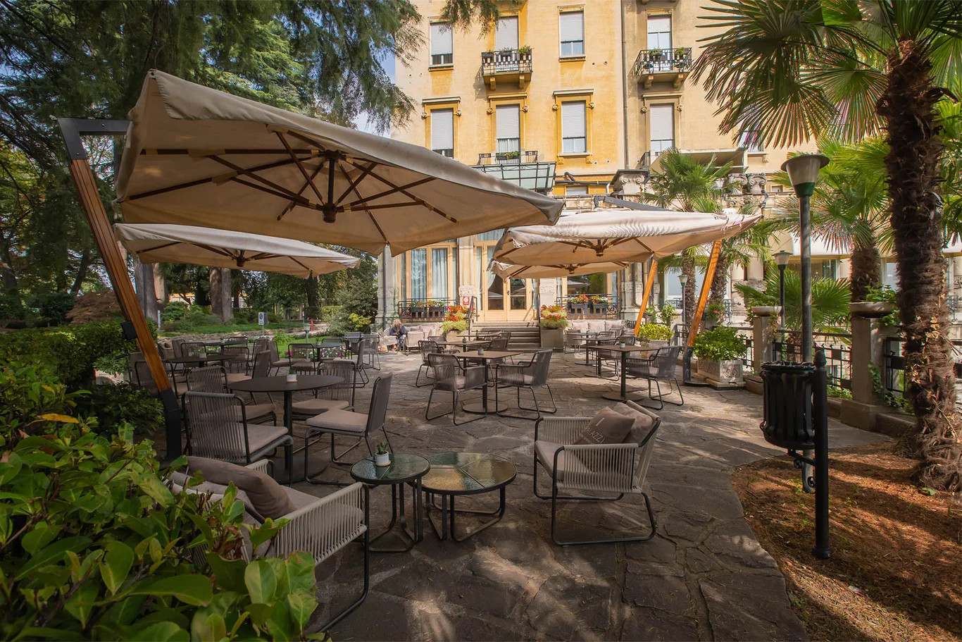 Eine elegante Terrasse mit Sonnenschirmen und Tischen, umgeben von üppigen Pflanzen und einem historischen Gebäude.