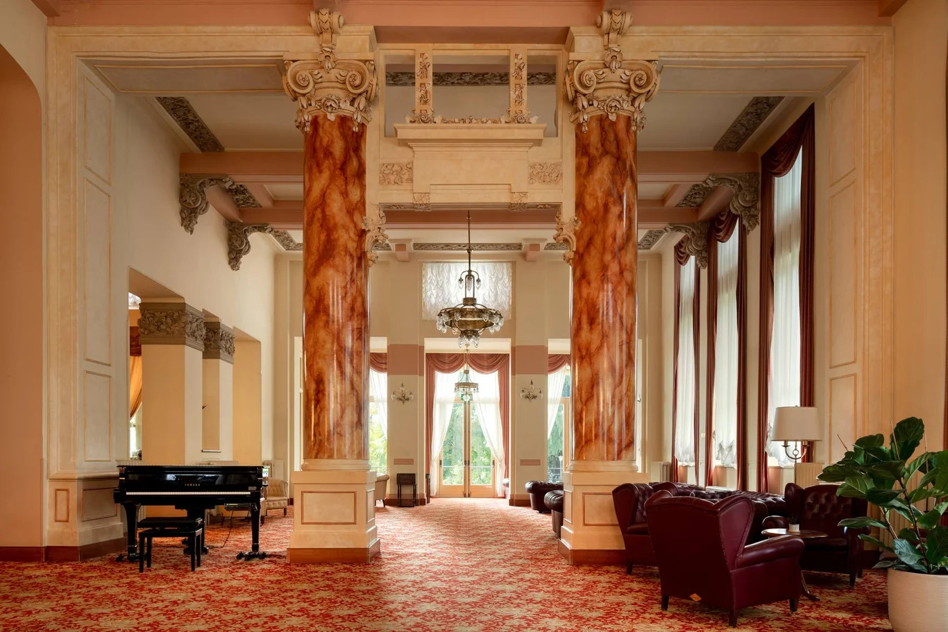 Ein eleganter Raum mit Säulen, rotem Teppich, einem Klavier und Blick auf eine Terrasse mit viel Tageslicht.