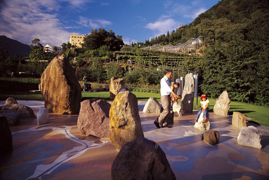 Eine Familie spielt auf einem bunten Platz mit Steinen, umgeben von Natur und Bergen im Hintergrund.