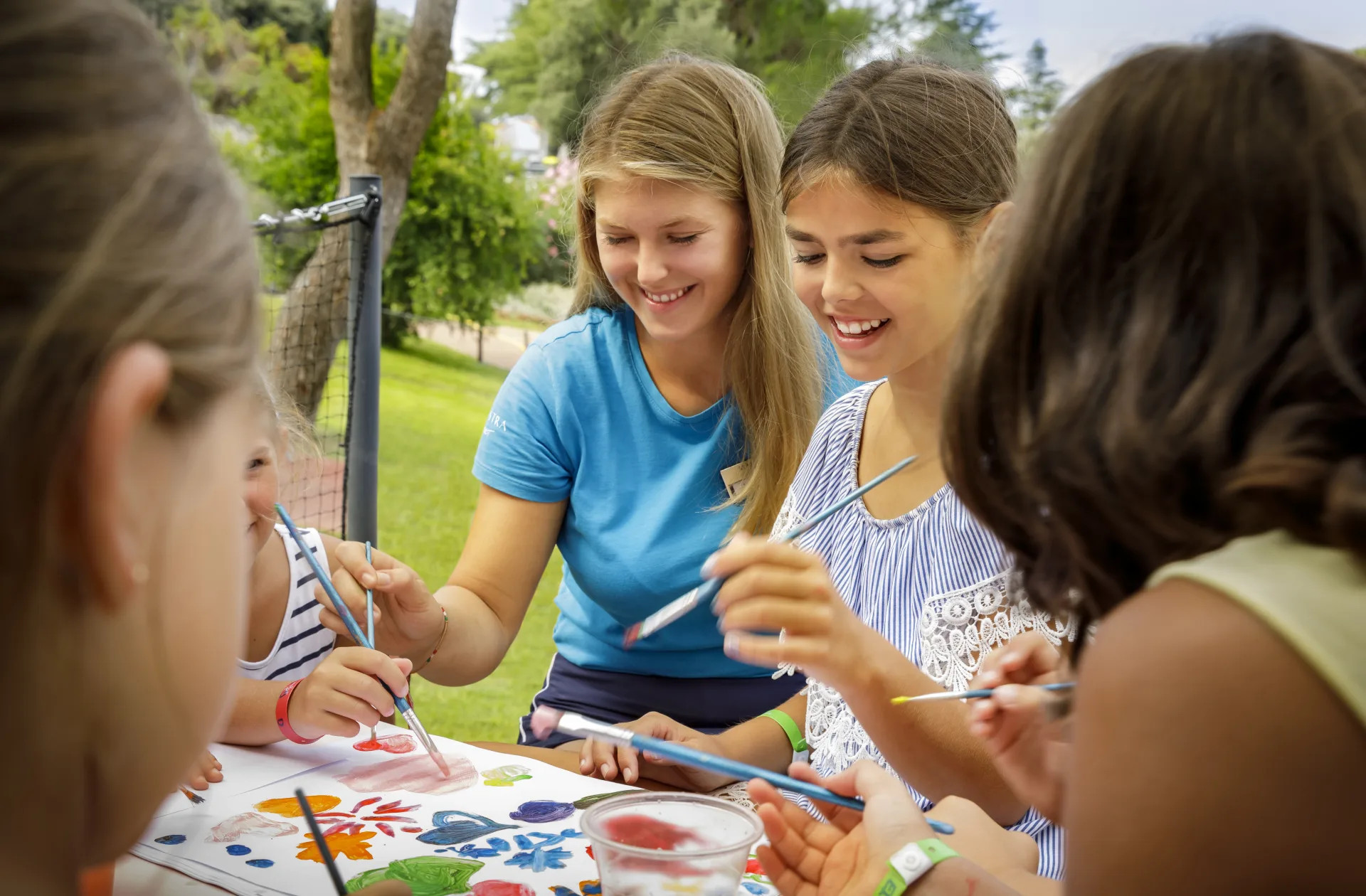 Kinder und eine Betreuerin malen fröhlich im Freien mit Pinsel und Farben an einem Tisch.