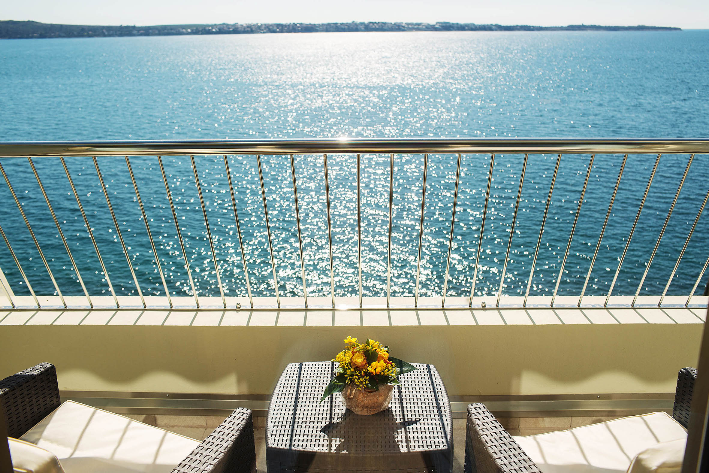 Balkon mit Aussicht auf glitzerndes Wasser und einem Blumenstrauß auf dem Tisch. Entspanntes Urlaubsambiente.