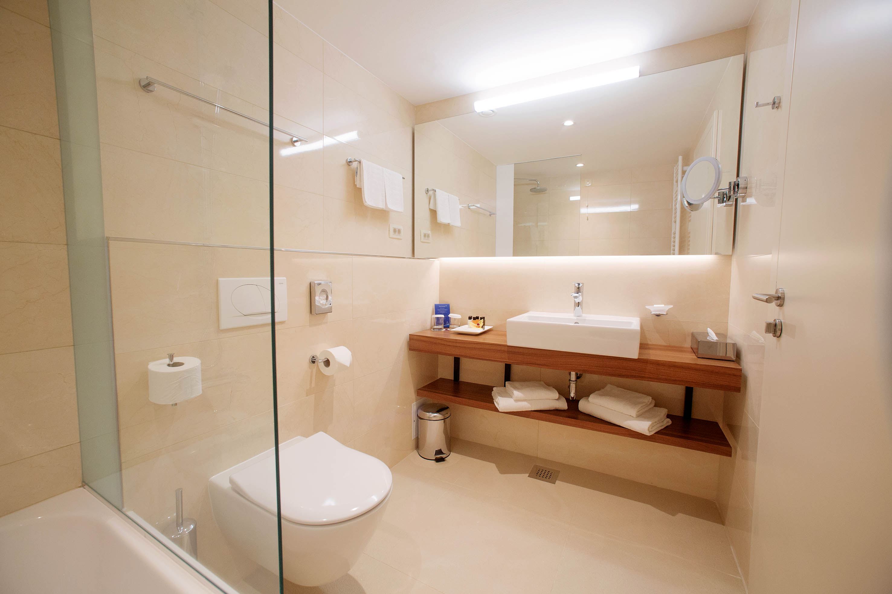 Modernes Badezimmer mit Dusche, Waschbecken und Holzregal. Helle Farben und minimalistische Ausstattung.