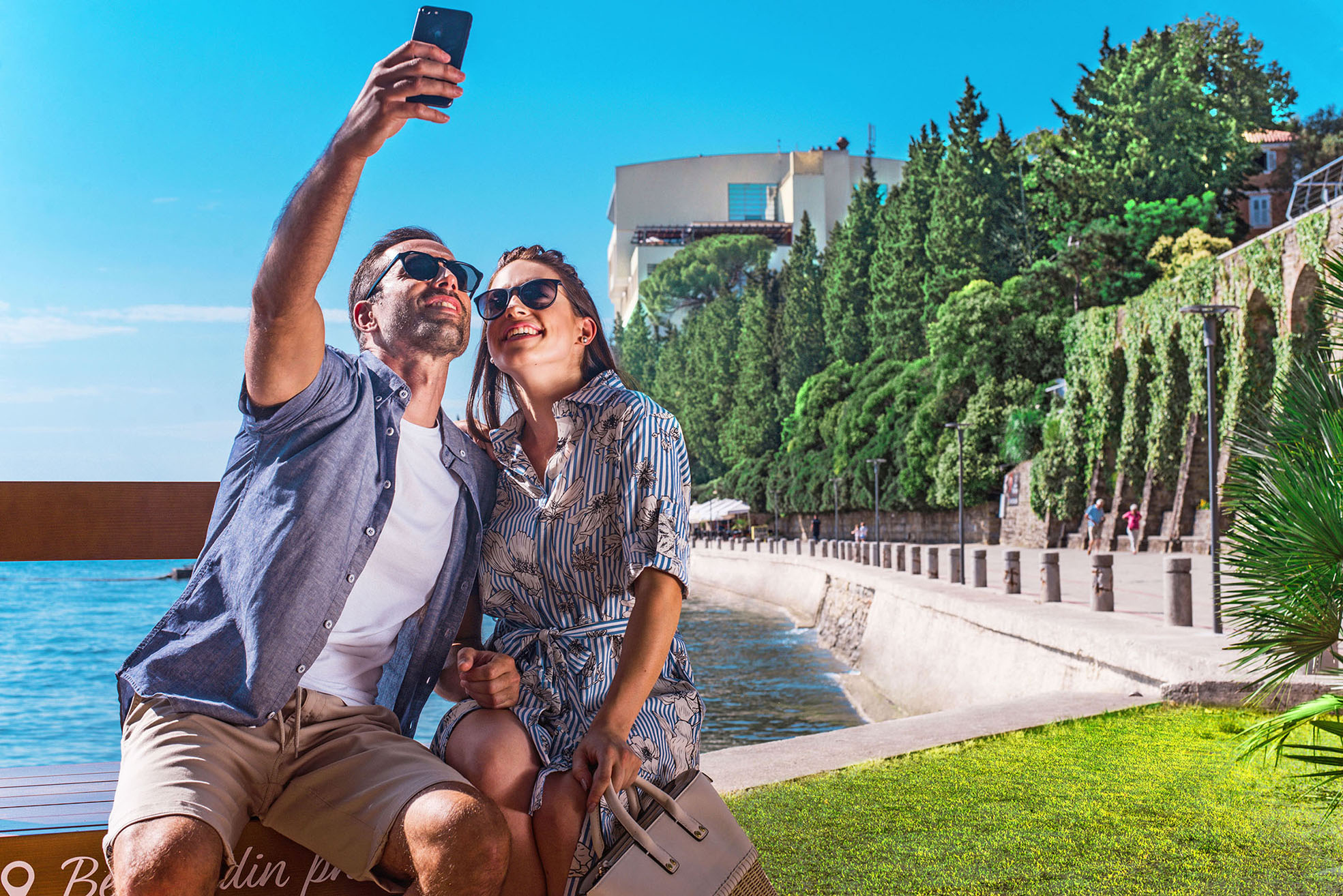 Ein Paar macht ein Selfie am Strand, umgeben von grünem Hintergrund und blauem Himmel. Sonne scheint.