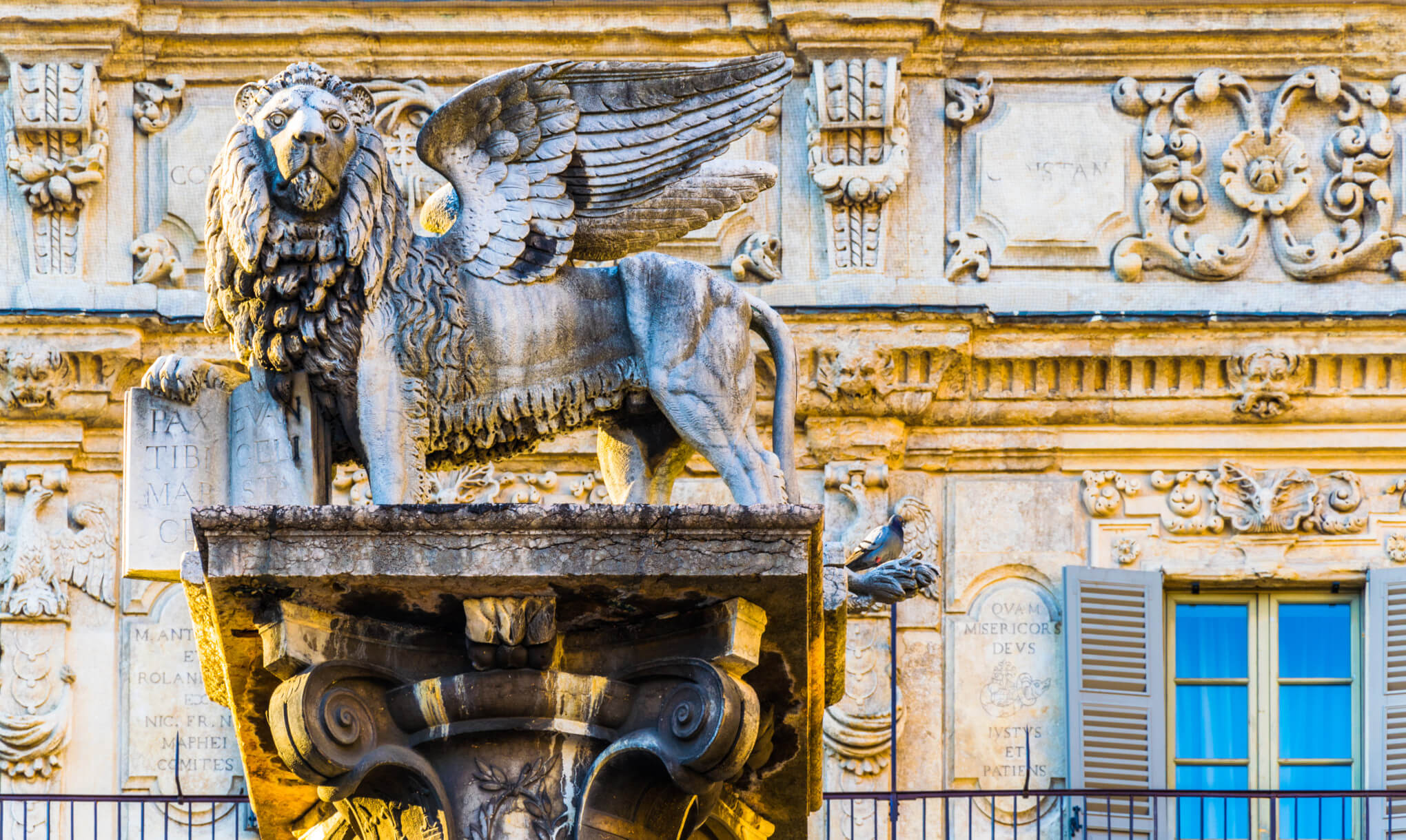 Ein geflügelter Löwe steht auf einem Säulenfundament, vor dekorierter Wand mit Fenstern.