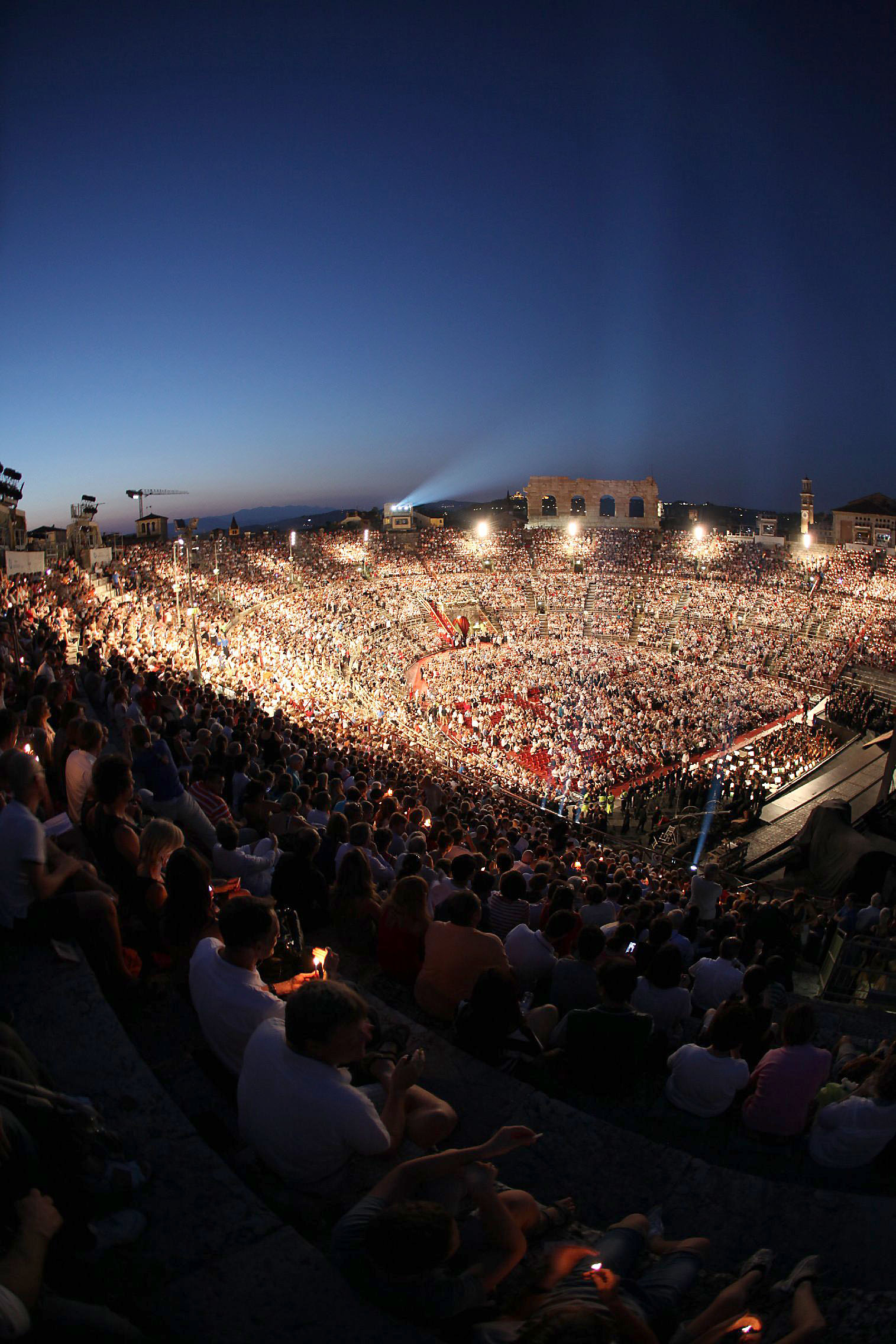 Ein großes Publikum in einem antiken Amphitheater, beleuchtet, bei Dämmerung. Atmosphäre voller Spannung und Erwartung.