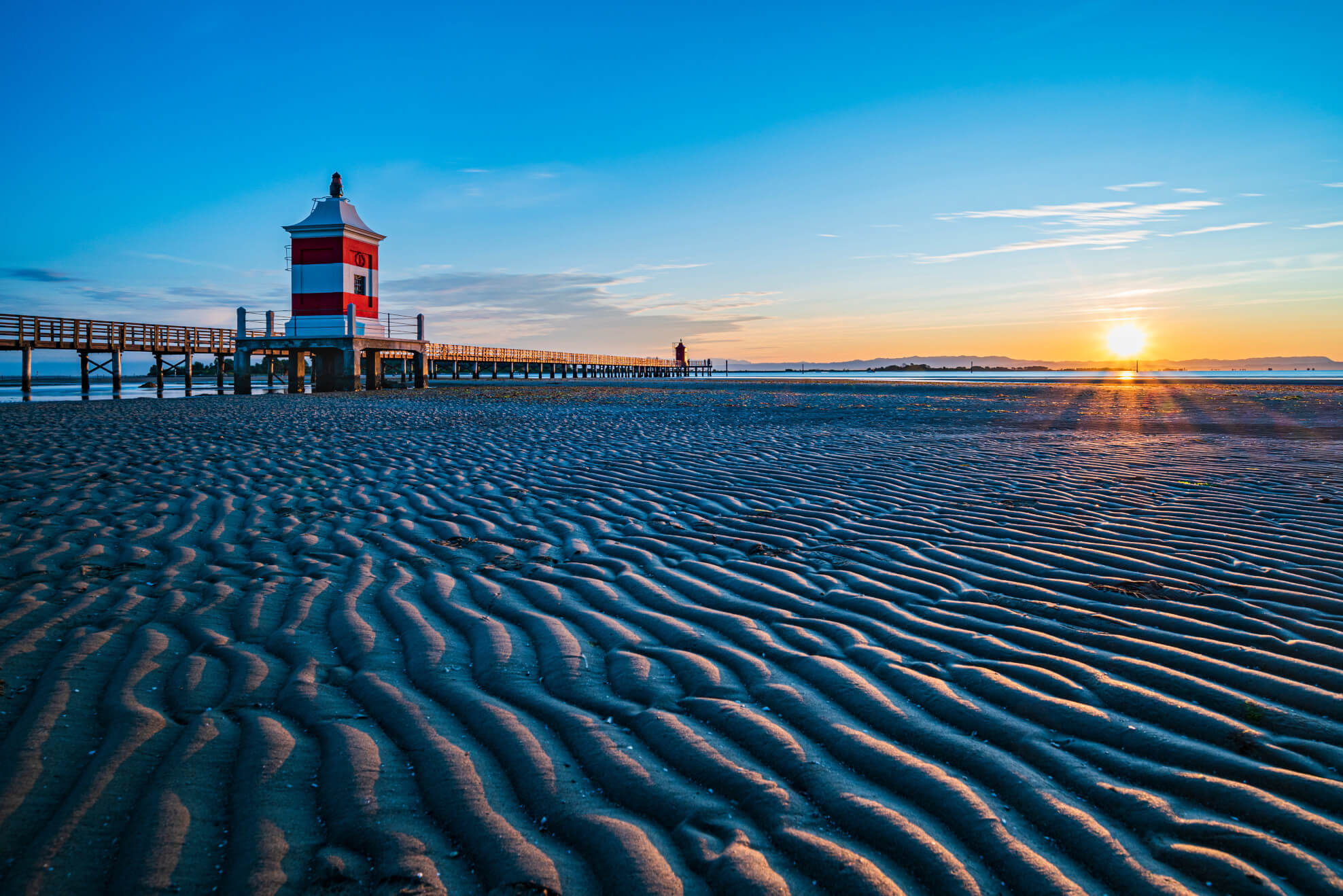 Ein Leuchtturm am Pier, bei Sonnenuntergang über einem gestreiften Sandstrand, in sanften Farbtönen.
