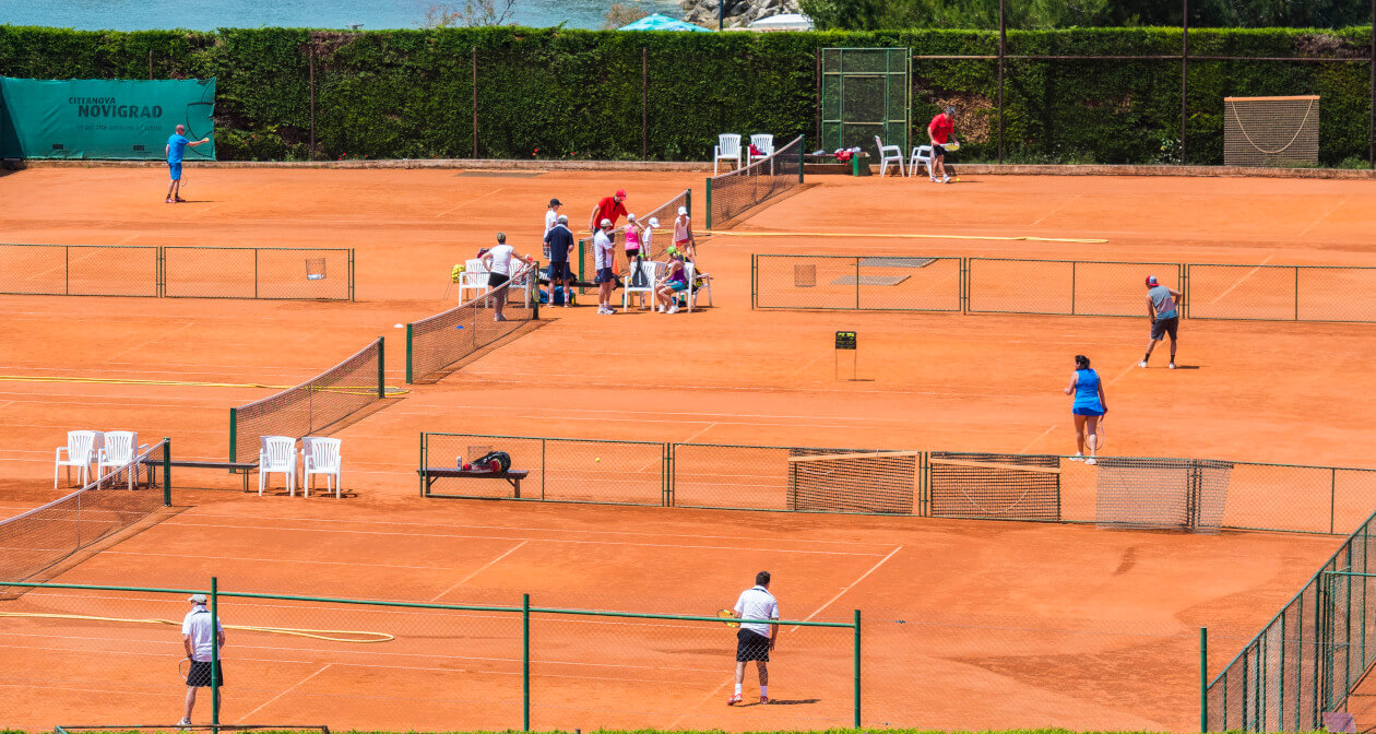 Tennisplatz mit mehreren Spielern, Zuschauern auf Stühlen und einer schönen Küstenlandschaft im Hintergrund.