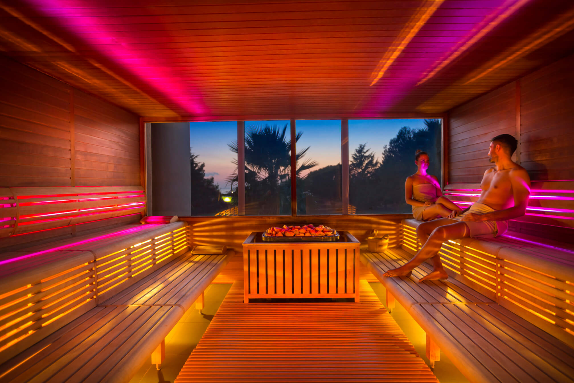 Ein modernes Saunainterior mit warmem Licht, zwei entspannenden Personen und Blick auf die Natur bei Sonnenuntergang.