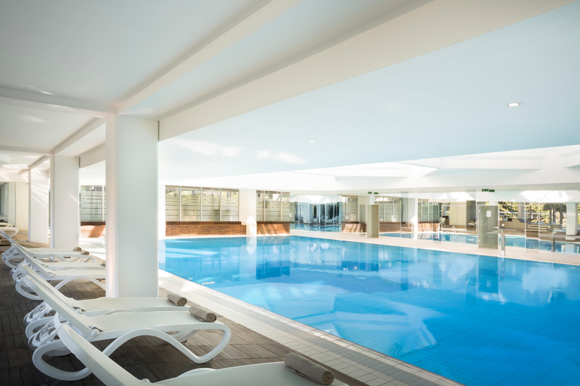 Ein modernes Hallenbad mit blauen Wasser und Liegen, umgeben von hellen Wänden und großen Fenstern.