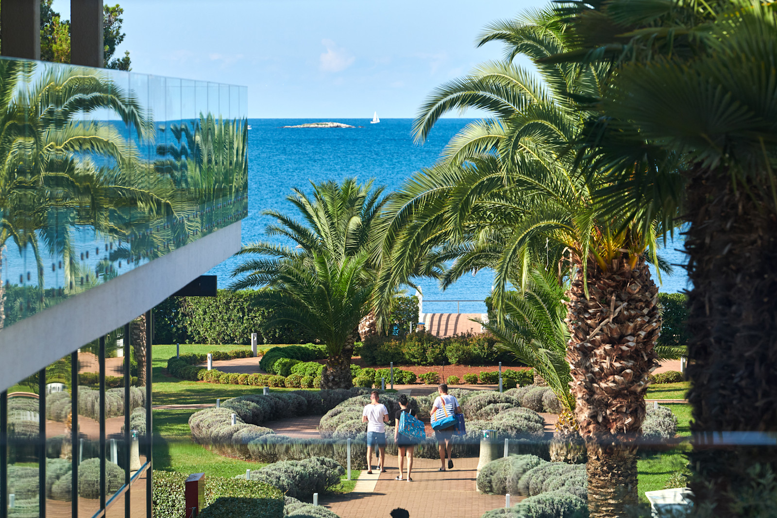 Drei Personen gehen an Palmen und einem blauen Meer entlang, mit einem modernen Gebäude im Hintergrund.