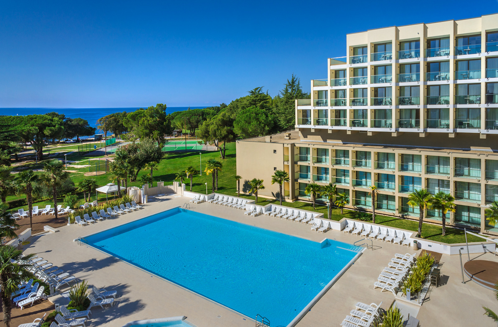 Ein sonniger Hotelpool mit Liegen, umgeben von Palmen und Blick auf das Meer und einen grünen Park.