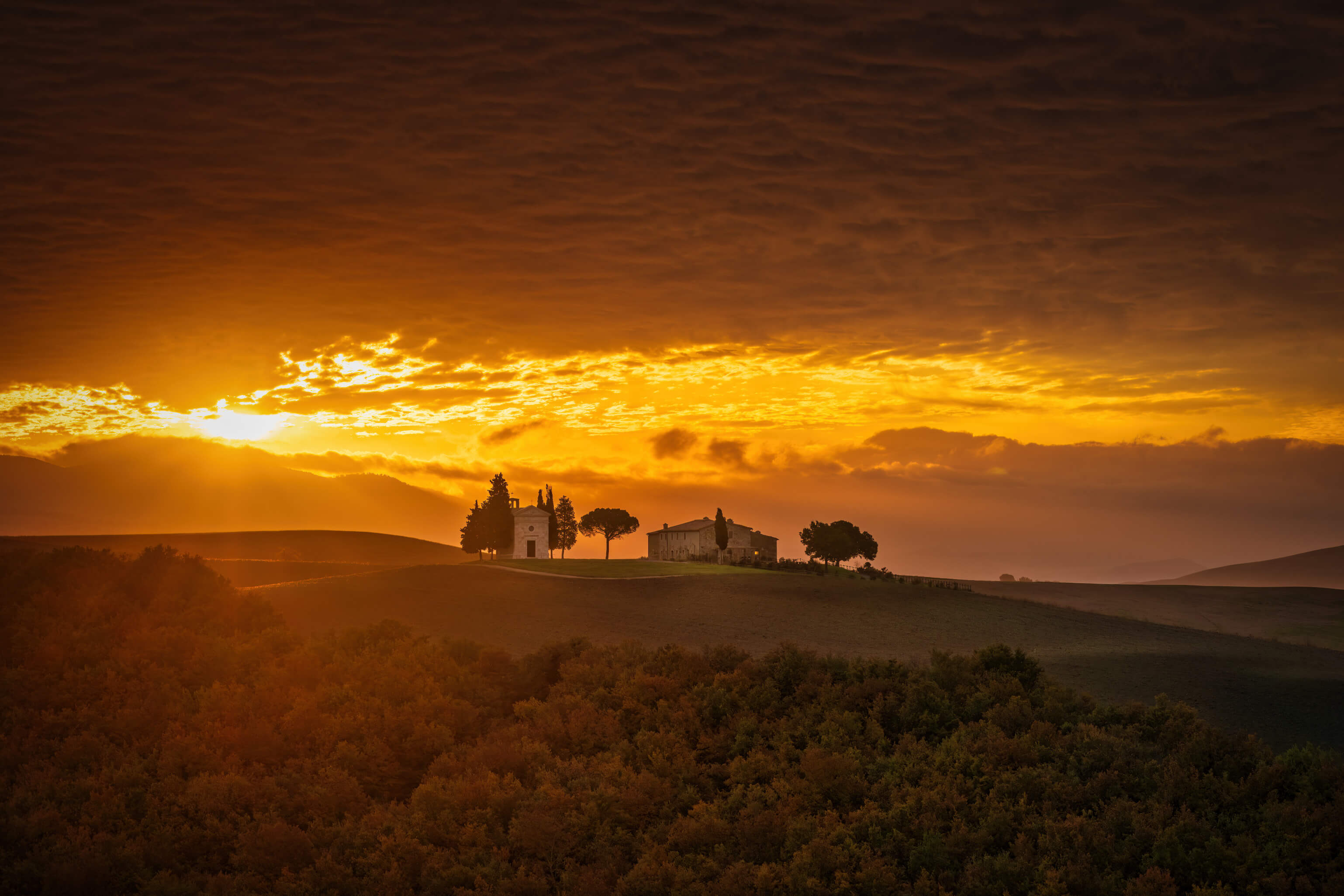 Eine malerische Landschaft bei Sonnenuntergang mit Hügeln und charmanten Gebäuden, umgeben von sanften Wolken.
