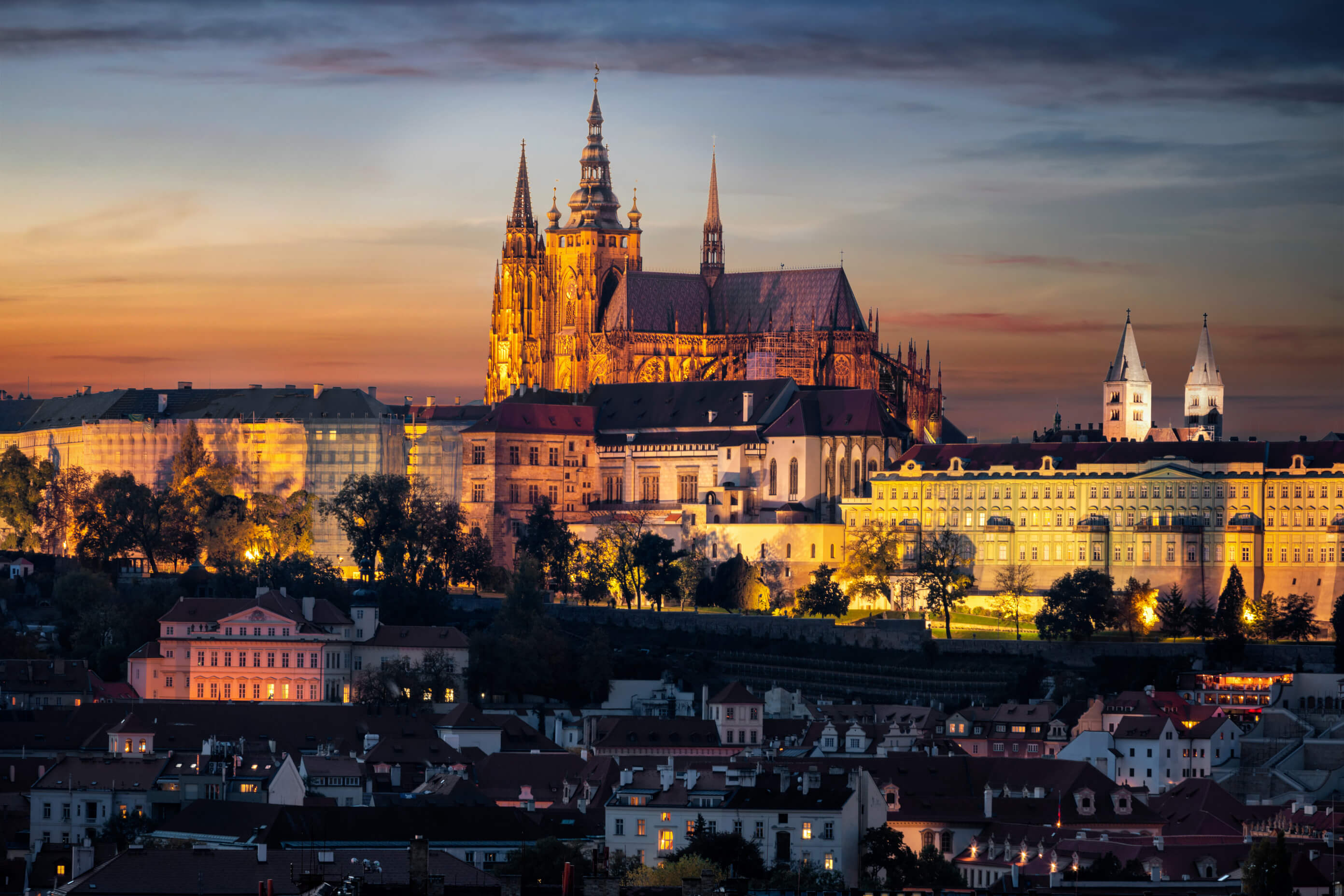 Die Prager Burg leuchtet majestätisch im Sonnenuntergang über der Stadt, umgeben von schönen Gebäuden und Bäumen.