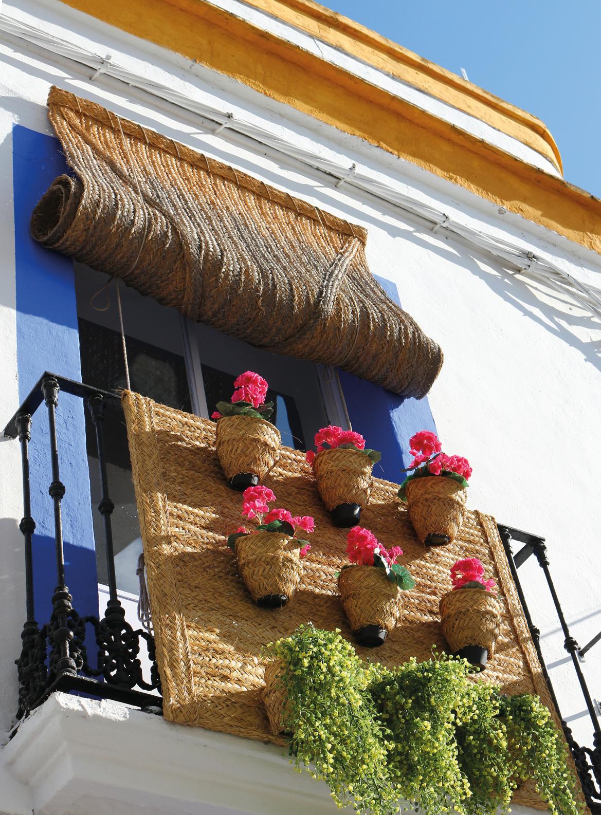 Ein Balkon mit geflochtenen Körben, roten Blumen und Pflanzen, vor weißer Wand und blauem Fensterrahmen.