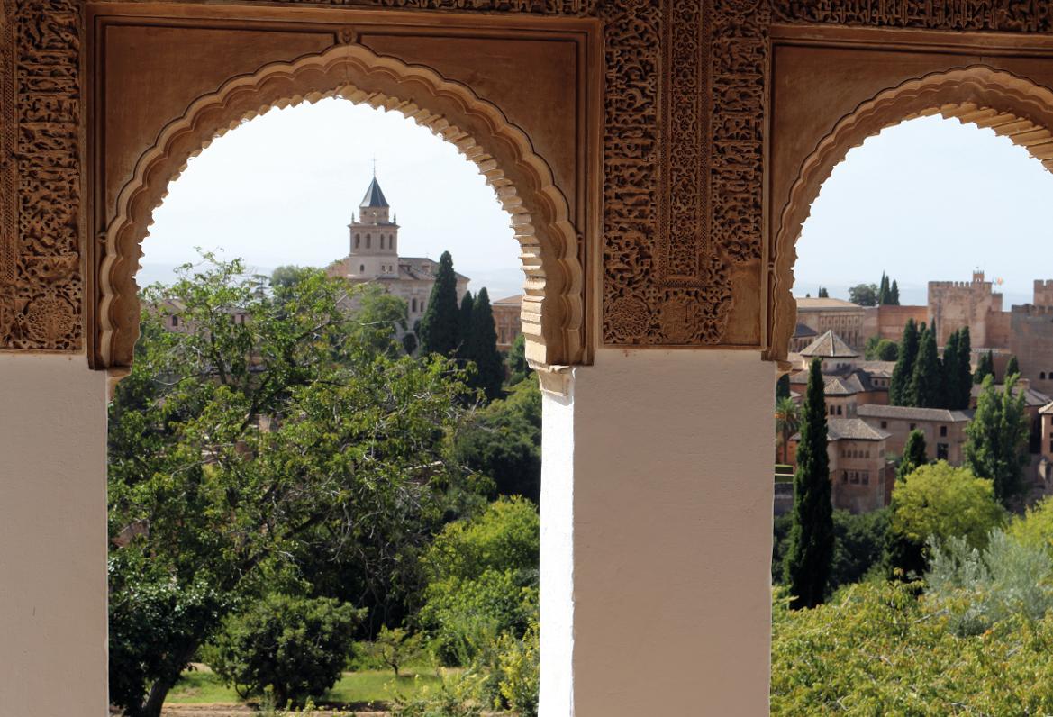 Blick durch ein kunstvoll verziertes Fenster auf die Alhambra und grüne Gärten im Hintergrund.