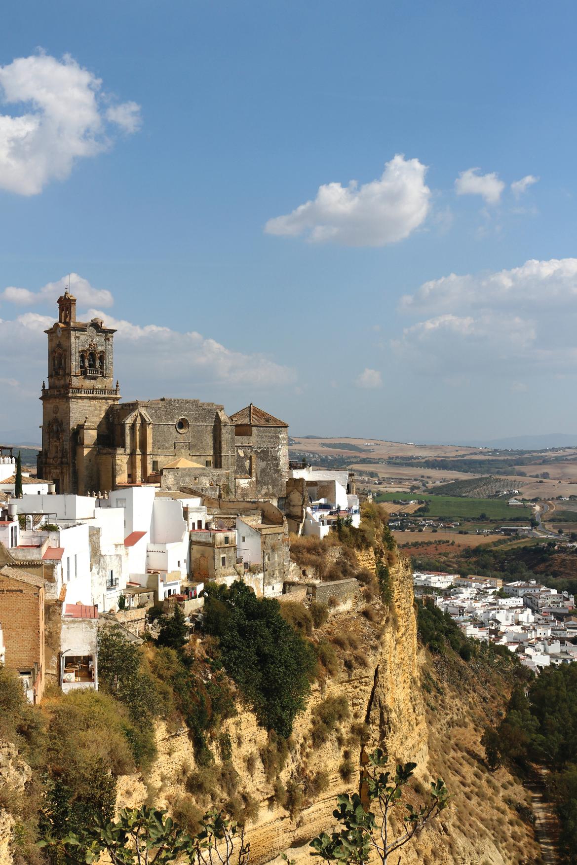 Eine historische Kirche thront über weißen Häusern auf einem Bergrücken mit hügeliger Landschaft im Hintergrund.