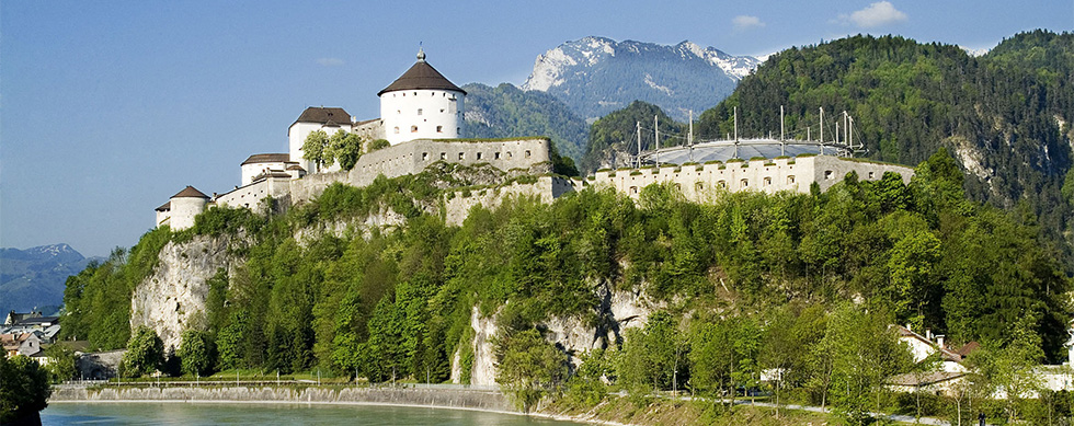 Eine beeindruckende Burg auf einem Hügel, umgeben von Bäumen und Bergen, unter blauem Himmel und hellem Licht.