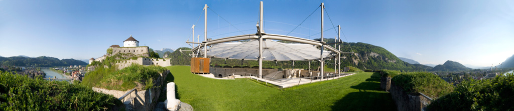 Eine panoramatische Aussicht auf eine Burg, umgeben von Bergen und einem Fluss, mit moderner Überdachung.