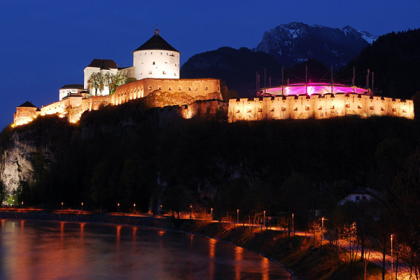 Eine beleuchtete Burg am Ufer eines Flusses, umgeben von Bergen, strahlt bei Nacht eine beeindruckende Stimmung aus.