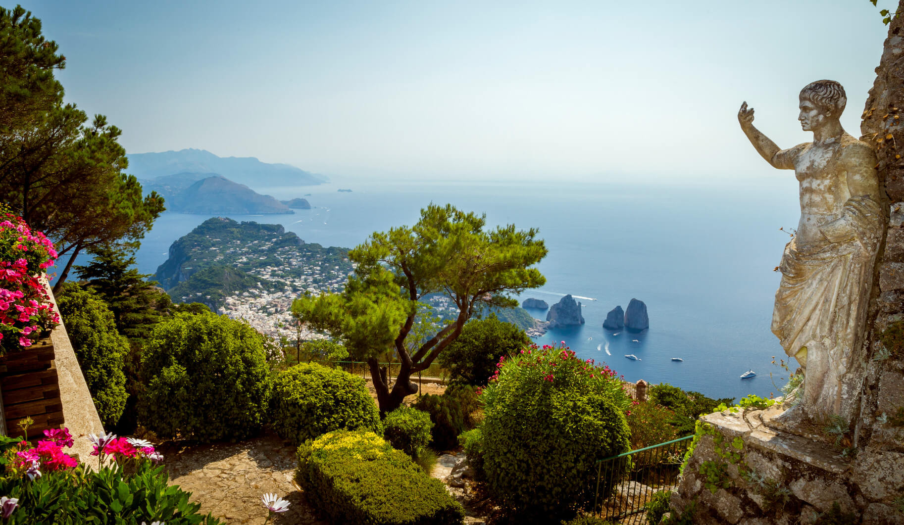 Blick über Capri mit bunten Blumen, einem Baum und einer Statue, die auf das Meer und die Felsen zeigt.