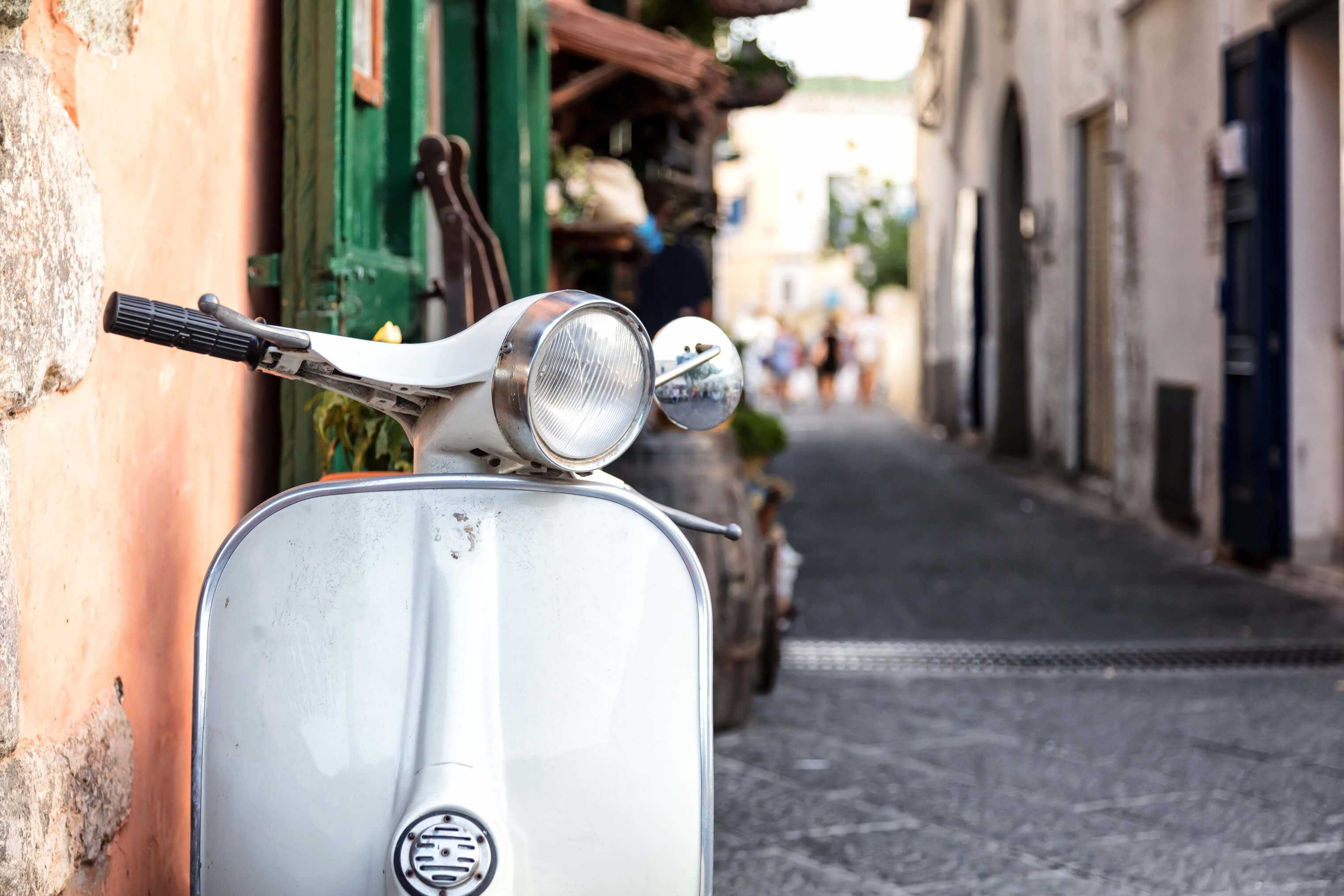 Eine weiße Vespa steht in einer schmalen Gasse, umgeben von charmanten alten Gebäuden und verschwommenen Passanten.