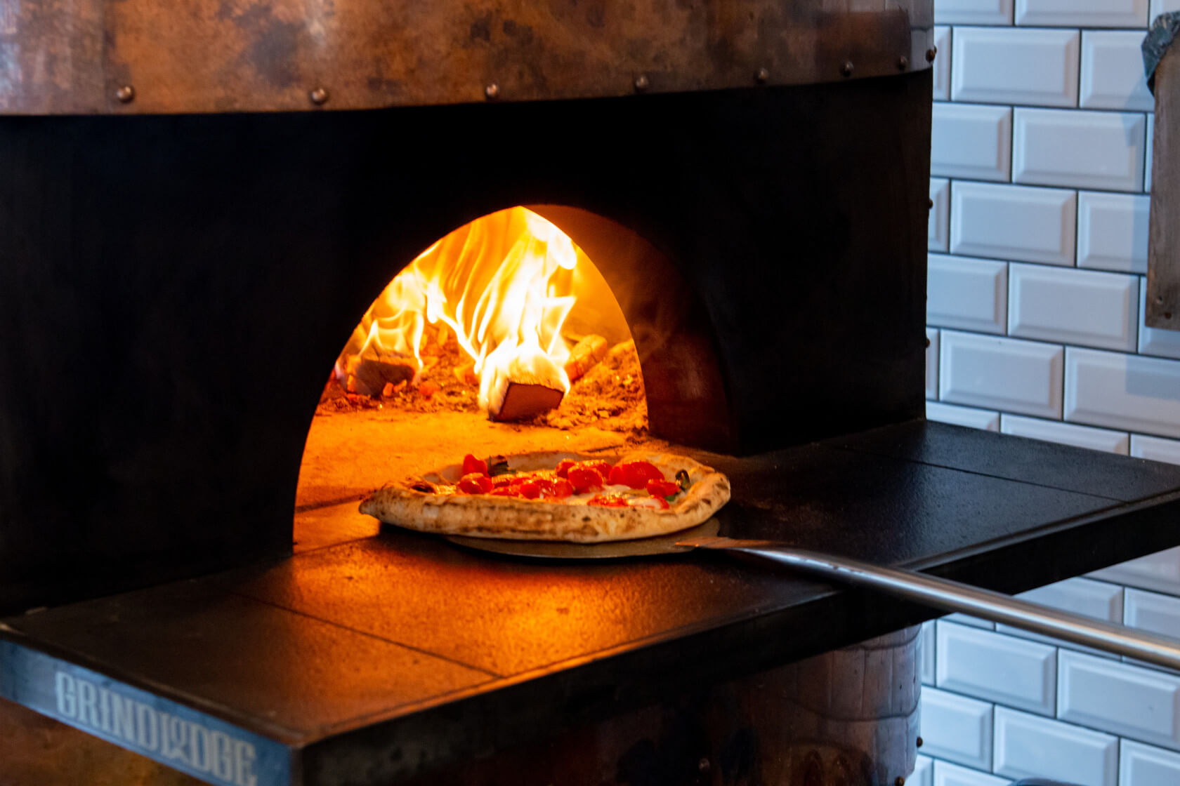 Ein Holzofen mit einer Pizza, die gerade gebacken wird, während Flammen sichtbar sind. Leckere Tomaten sind drauf.