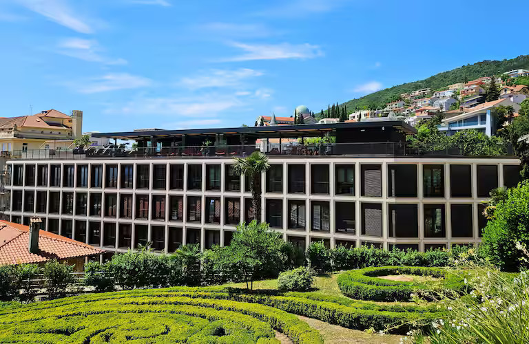 Ein modernes Gebäude mit Terrasse, umgeben von Gärten und Hügeln unter einem blauen Himmel.