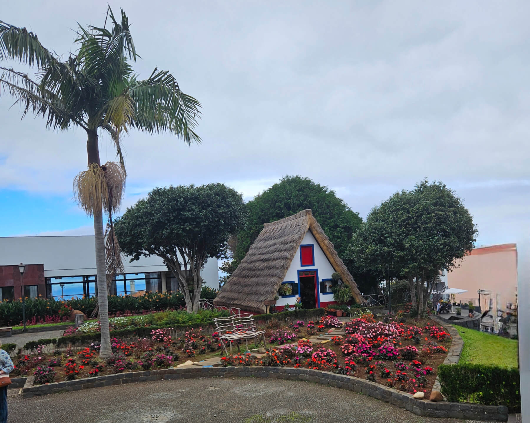 Ein buntes Blumengarten mit einem strohgedeckten Häuschen und Palmen im Hintergrund. Wolkiger Himmel darüber.