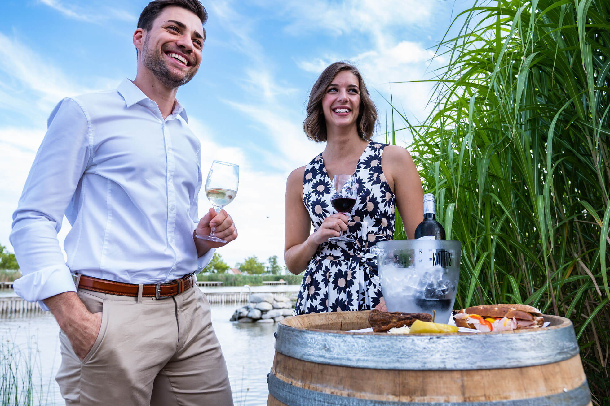 Ein Paar genießt Wein und Snacks an einem Teich, umgeben von üppigem Grün und einem klaren Himmel.