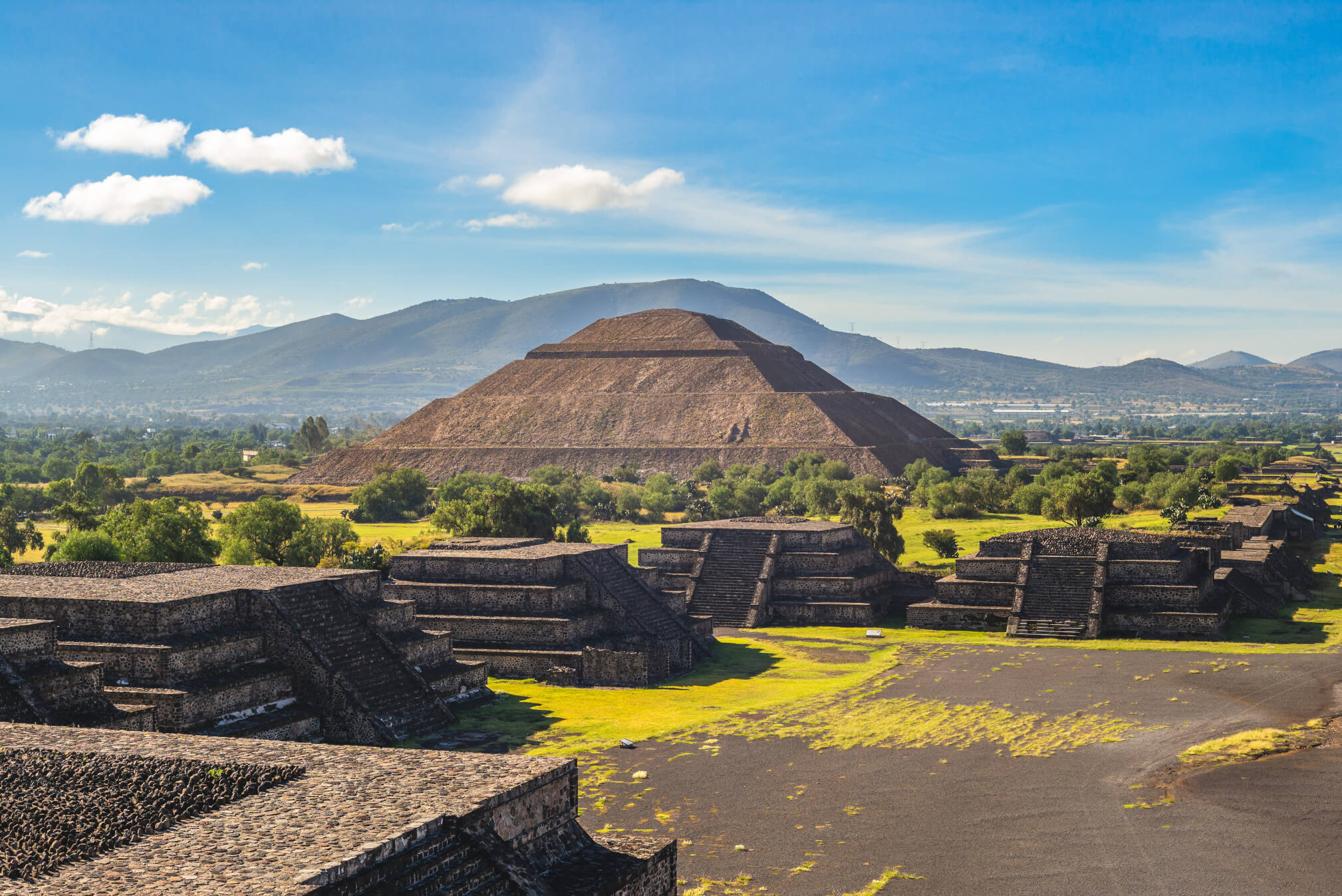 Die Pyramide von Teotihuacan erhebt sich majestätisch mit grünen Flächen und Bergen im Hintergrund. Schöner Himmel!