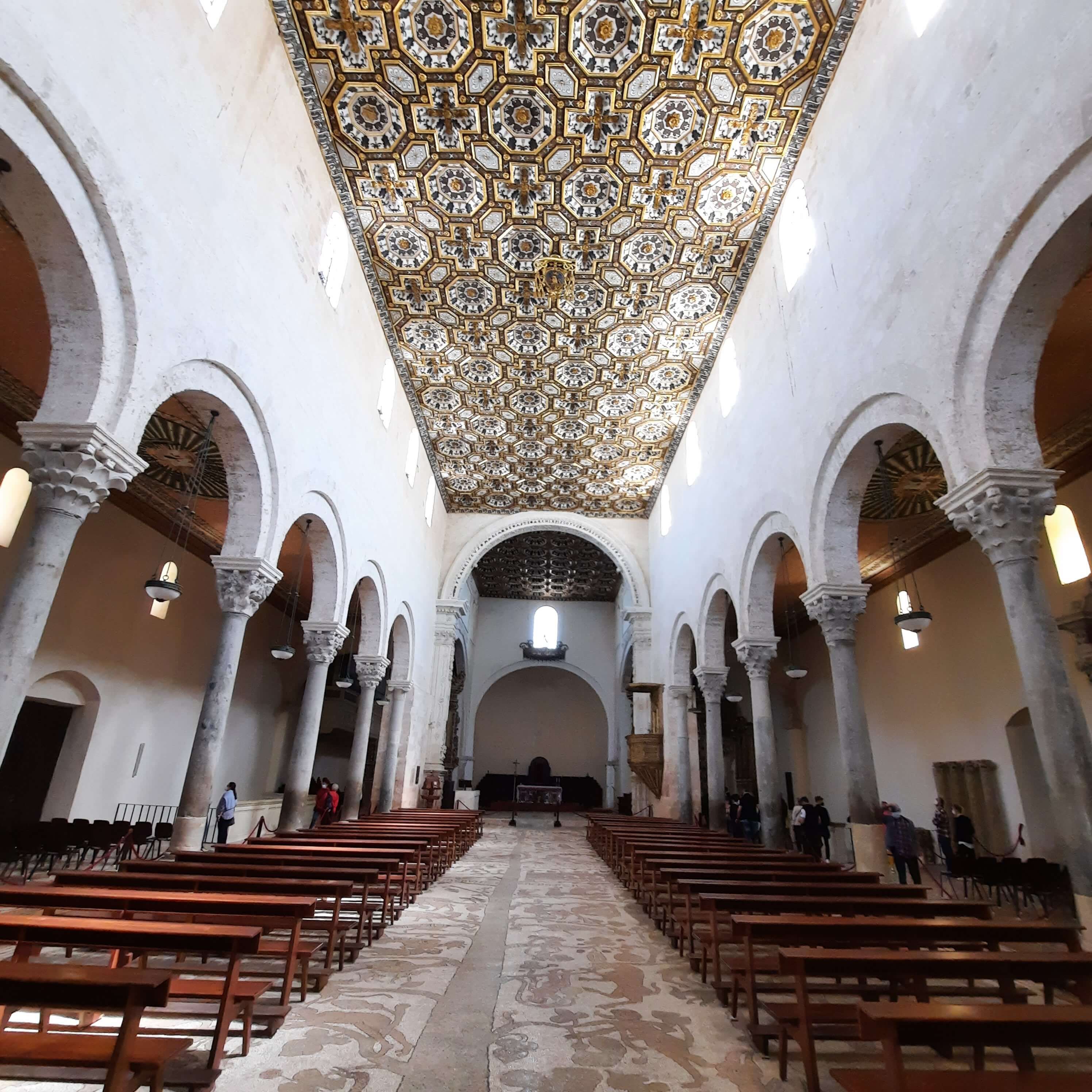 Eine beeindruckende Kirche mit hohen Säulen und einem kunstvollen, goldenen Deckendesign. Leere Bänke laden zur Ruhe ein.