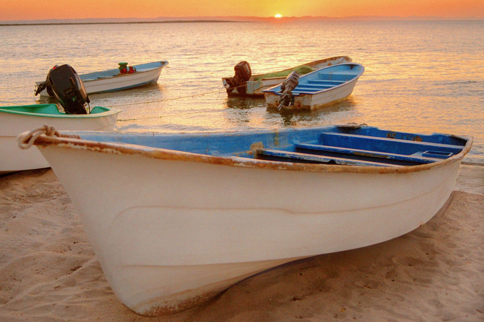 Fünf Boote stehen am Ufer, während die Sonne über dem Wasser untergeht und den Himmel in warmen Farben färbt.