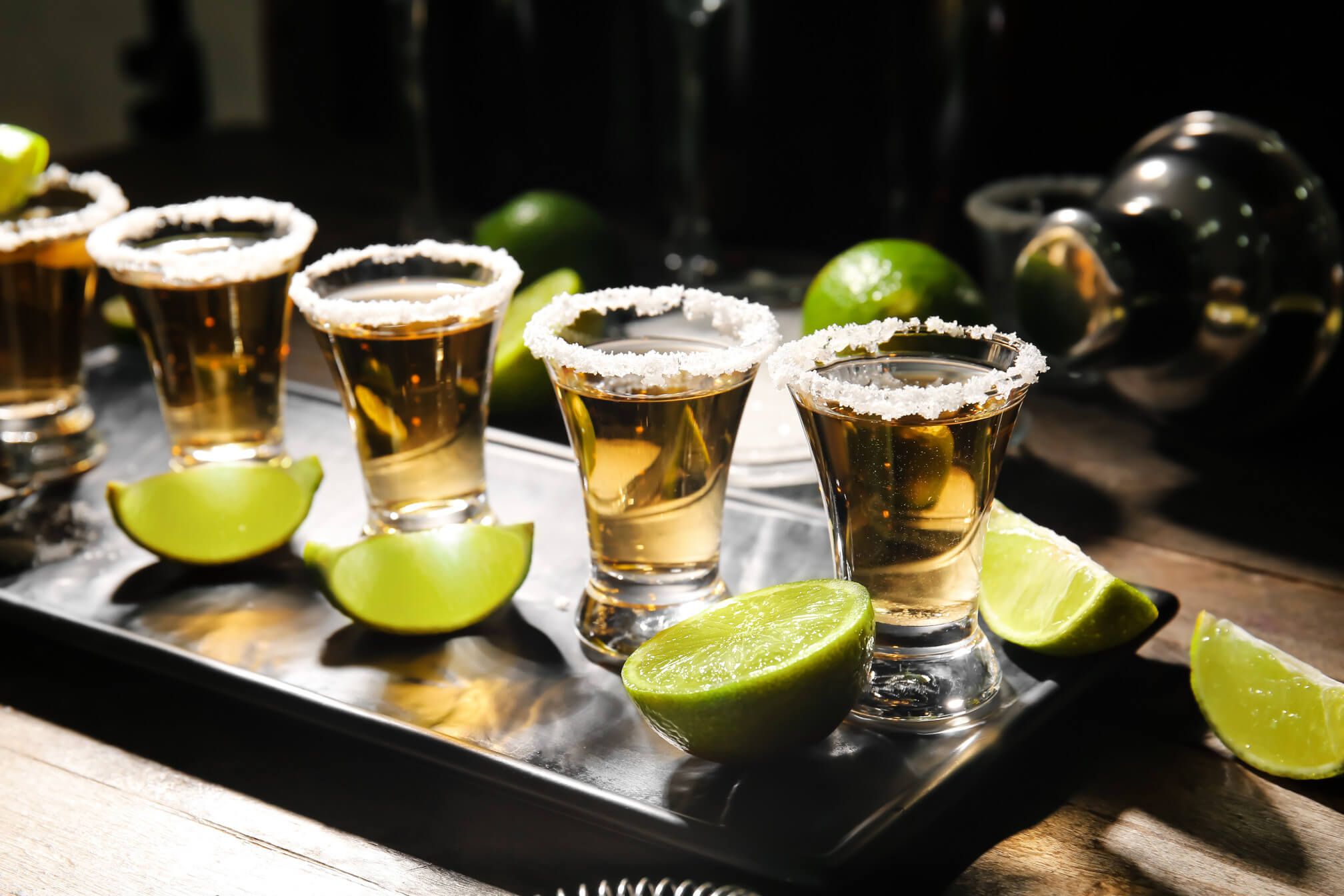 Fünf Tequila-Shots mit Salzrand und Limettenscheiben auf einem schwarzen Tablett. Atmosphäre ist festlich.