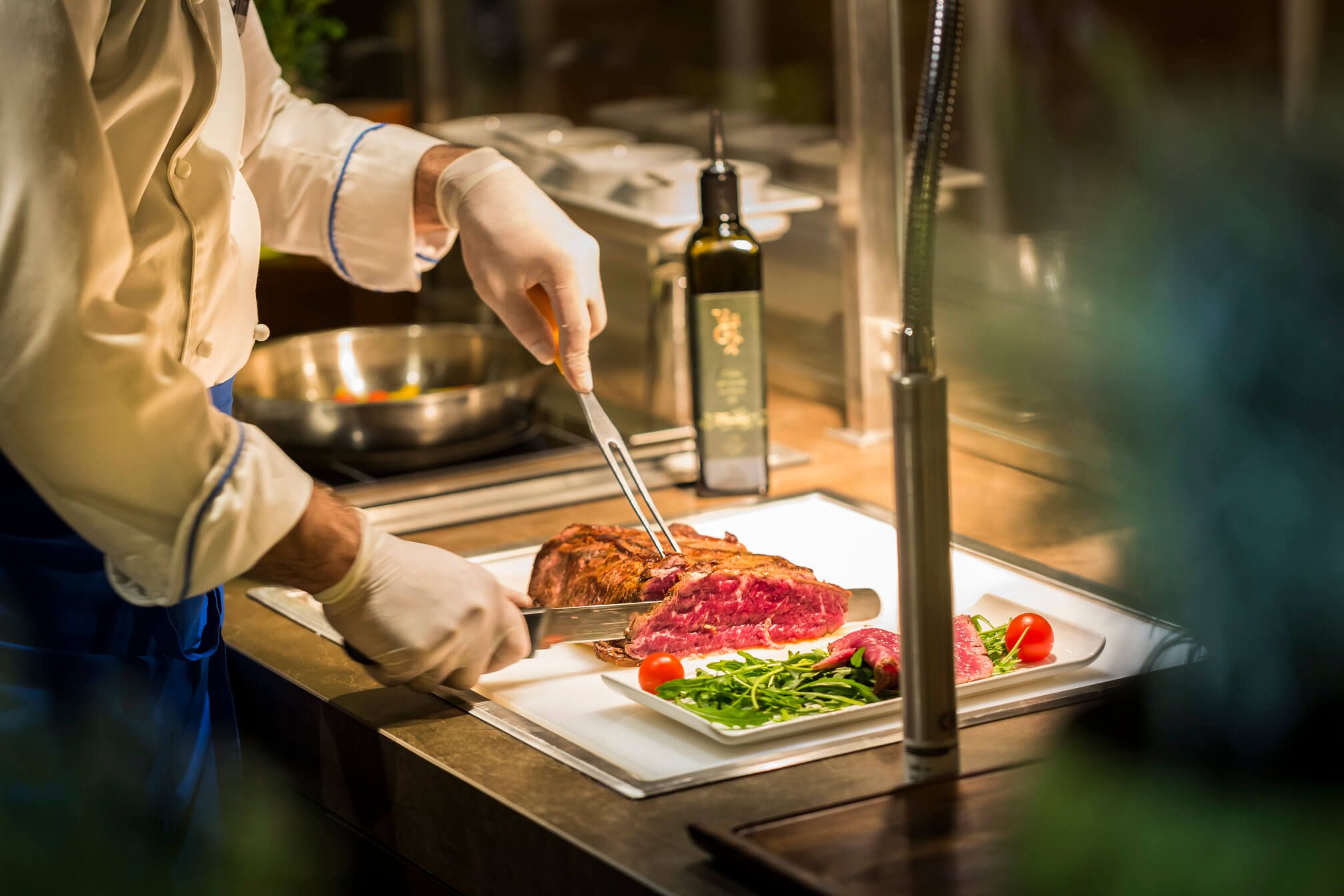 Ein Koch schneidet saftiges Fleisch auf einem Teller, umgeben von frischem Gemüse und Kräutern.