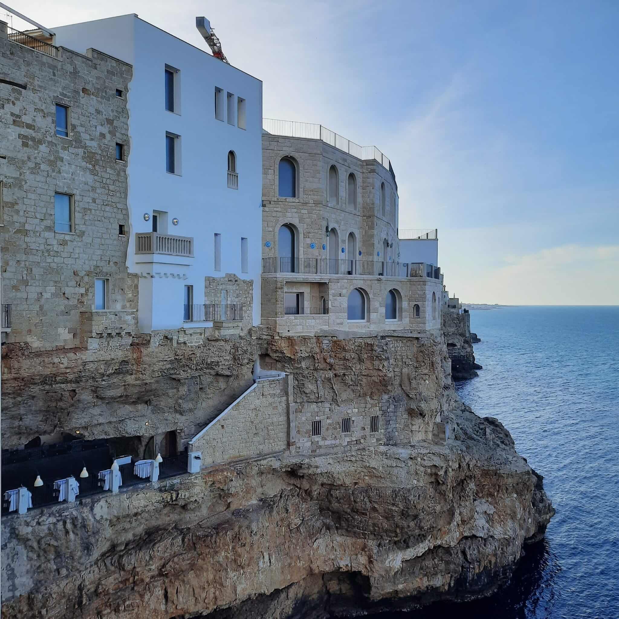 Felsen und historische Gebäude am Meer, mit blauer Wasseroberfläche im Hintergrund und klarem Himmel.