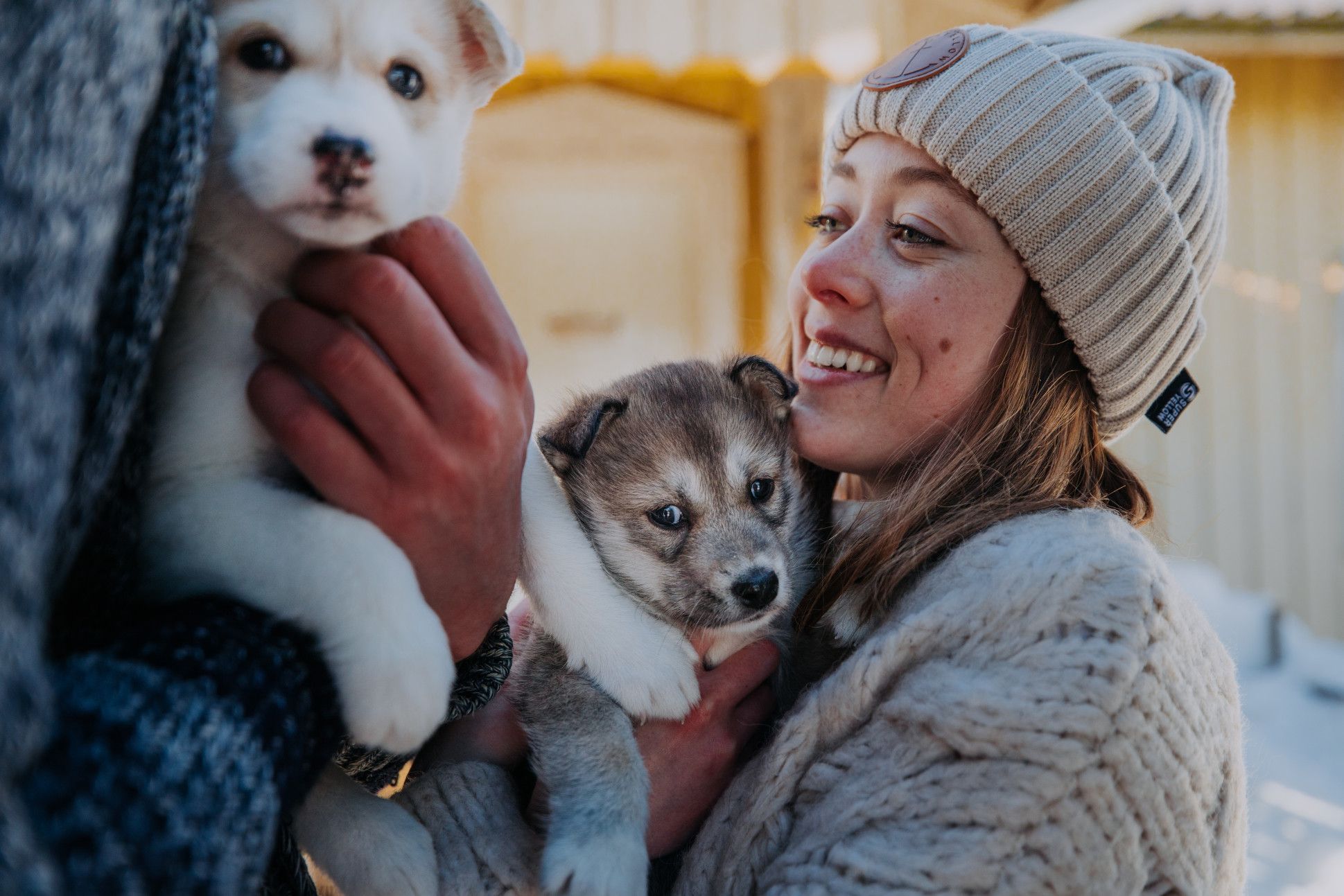 Eine Frau lächelt, während sie zwei süße Welpen in ihren Armen hält. Es ist ein fröhlicher, winterlicher Moment.