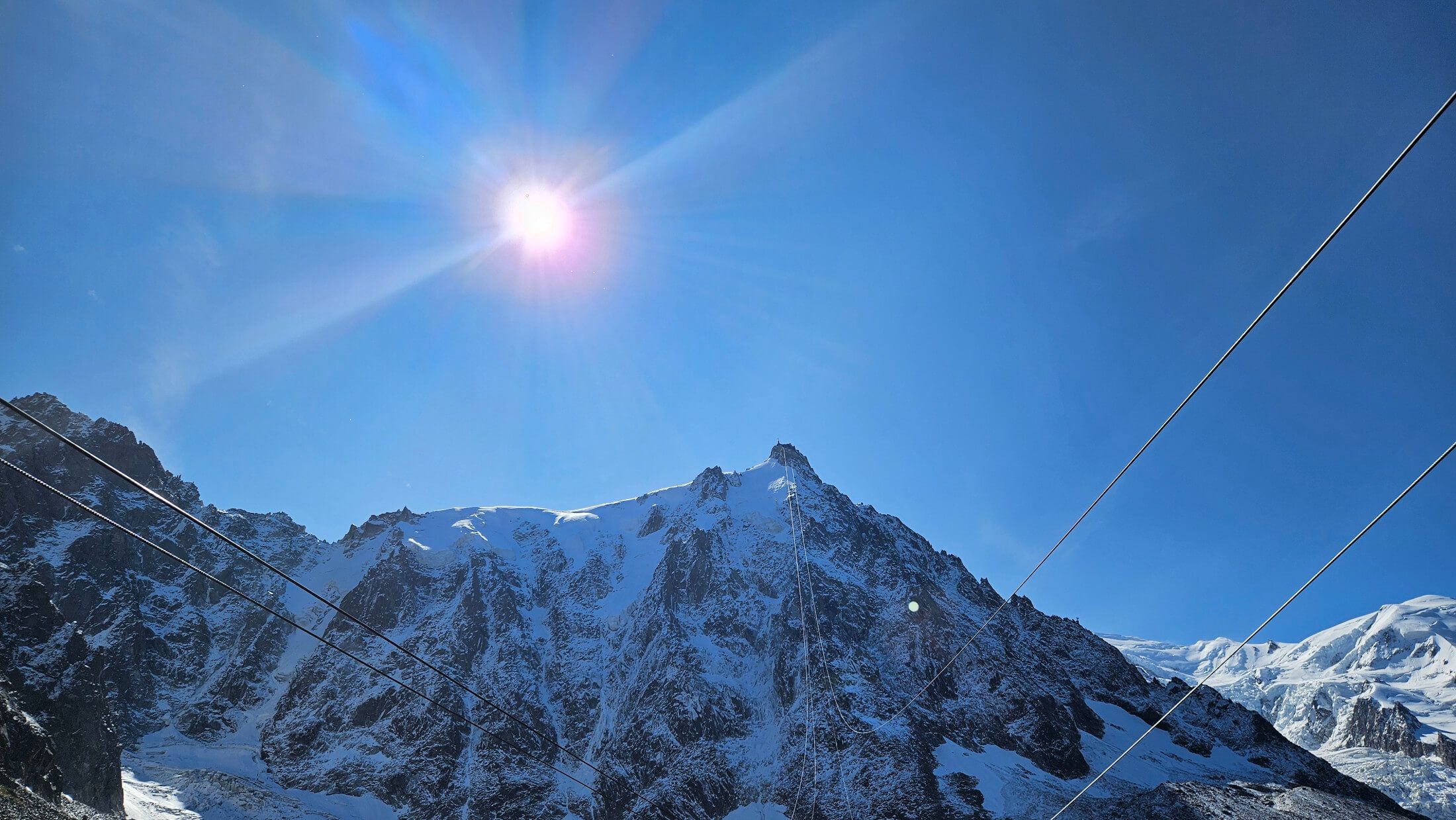 Sonniger Himmel über schneebedeckten Bergen, mit Seilbahnkabeln, die die majestätische Landschaft durchziehen.
