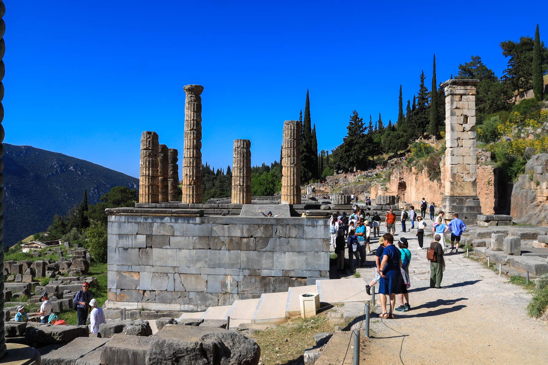 Eine antike Stätte mit Säulen, umgeben von Besuchern, unter klarem blauen Himmel und Bergen im Hintergrund.