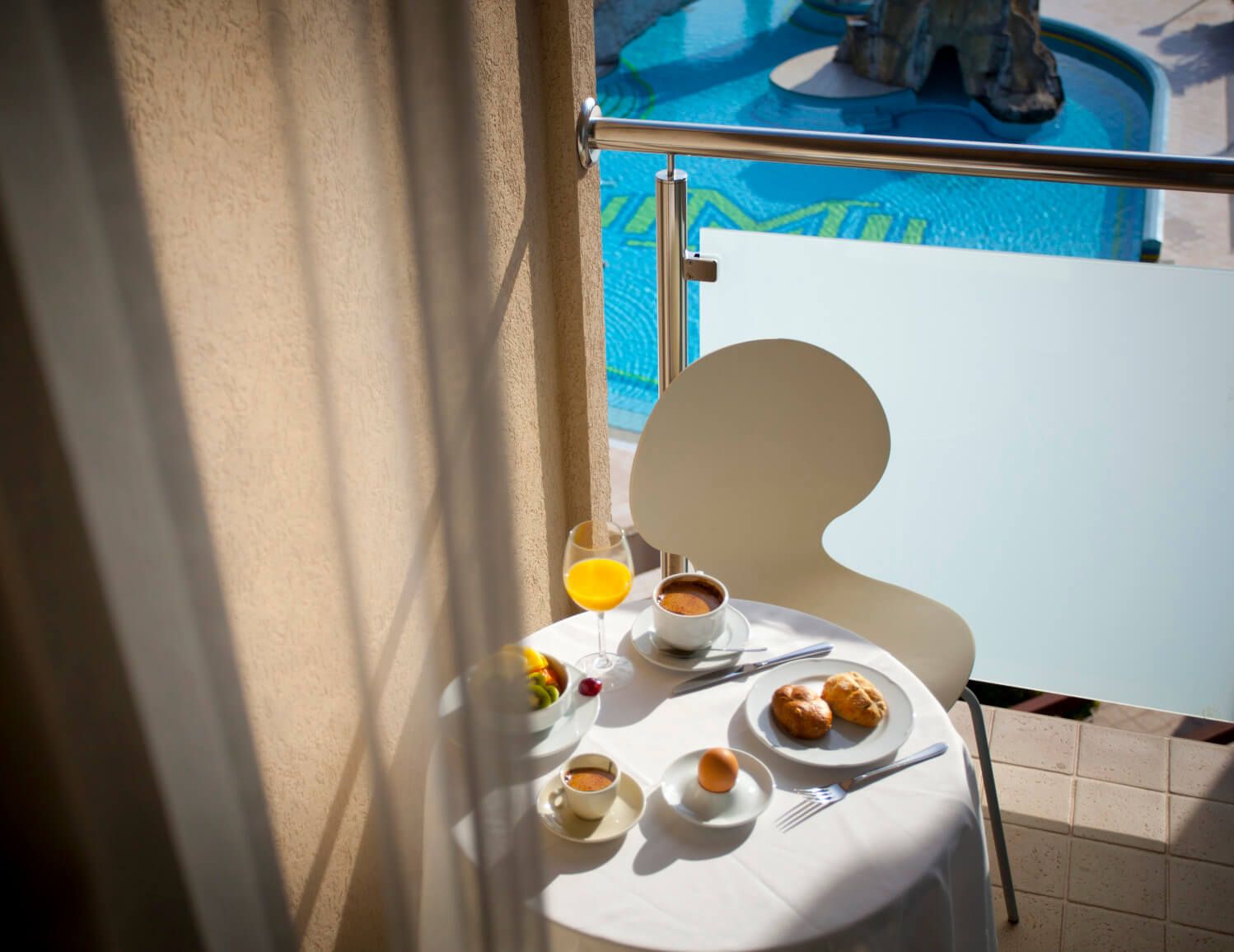 Ein Frühstückstisch auf einem Balkon mit Blick auf einen blauen Pool, umgeben von frischem Obst und Getränken.