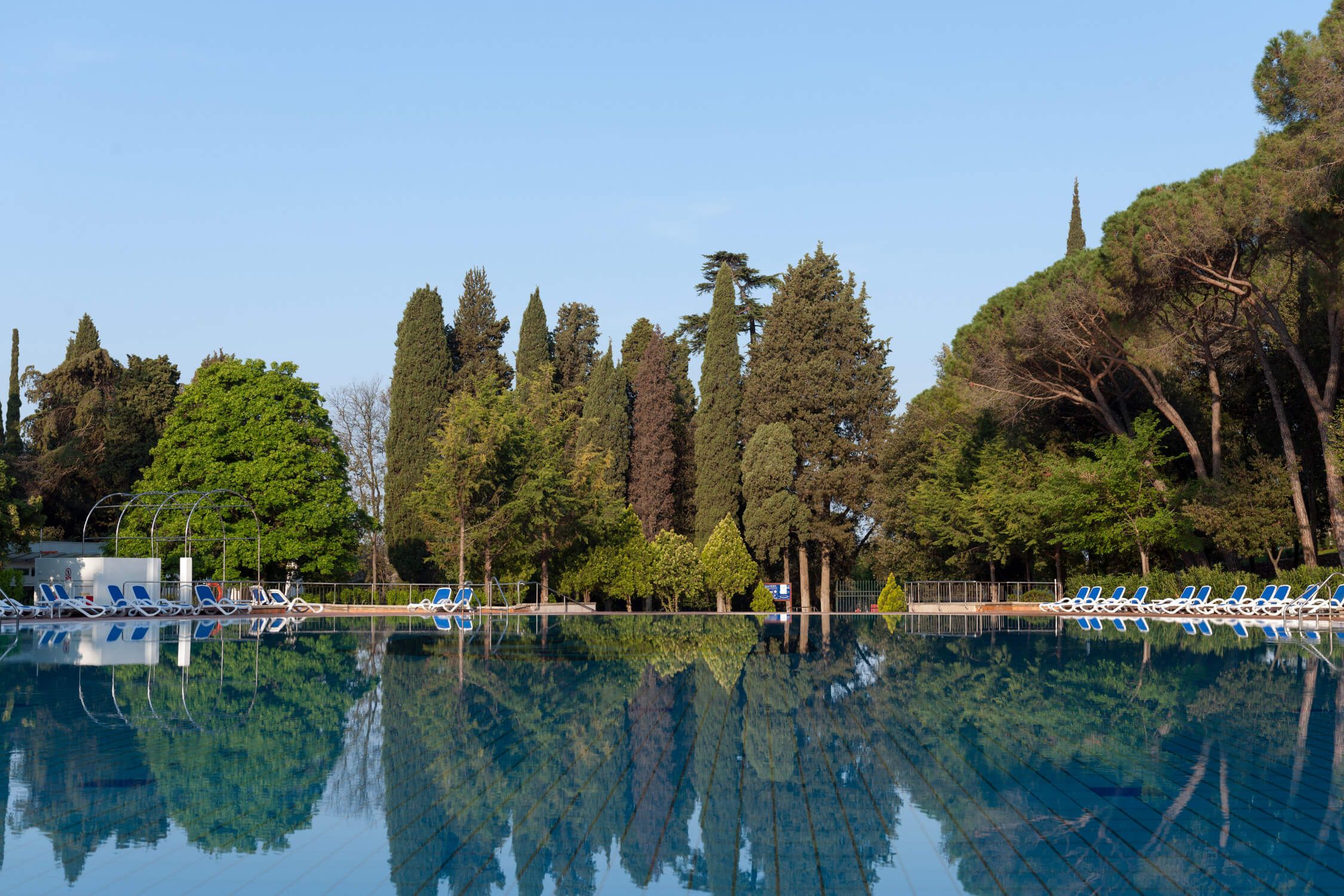 Ein ruhiger Pool mit Spiegelung der Bäume unter blauem Himmel, wenige Liegen am Rand.