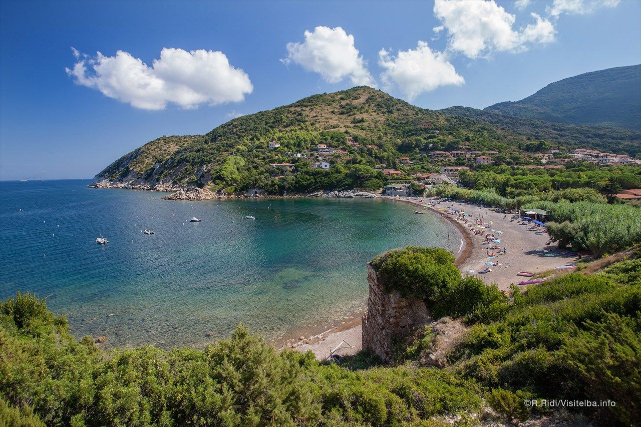 Eine malerische Bucht auf Elba mit klarem Wasser, Stränden und umgebenden Hügeln unter blauem Himmel und weißen Wolken.